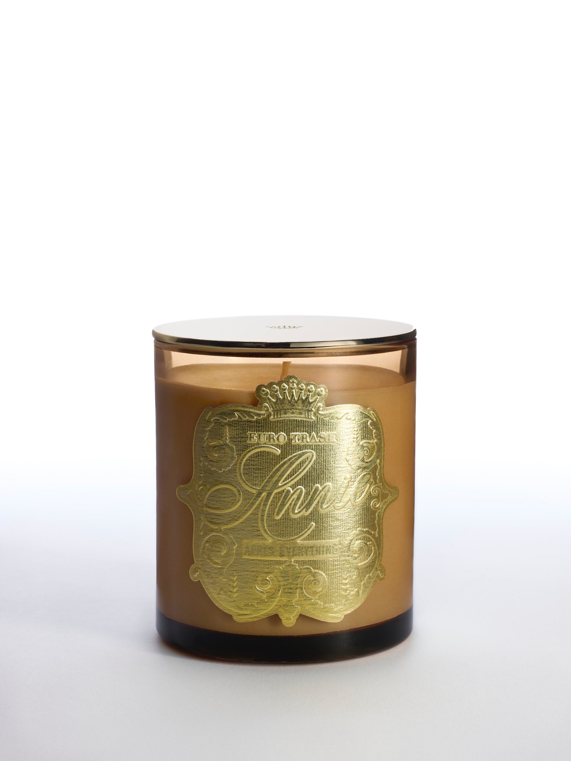 The Apres Everything brown glass amber scented candle features a shiny gold decorative label with ornate text and patterns, shown on a white background.