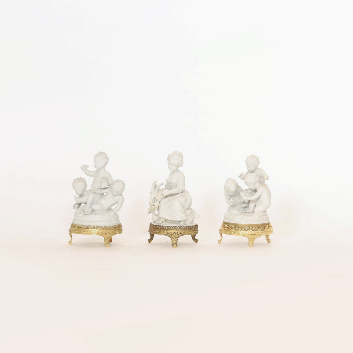 Displayed against a plain white background, the Set of Porcelain Cherub Figurines on Ormolu Bases by Euro Trash Annie features three elegant figures: a central woman flanked by groups of charming cherubic children. With ornate ormolu bases and classic porcelain detailing, these decorative collectibles bring an elegant touch to any display.