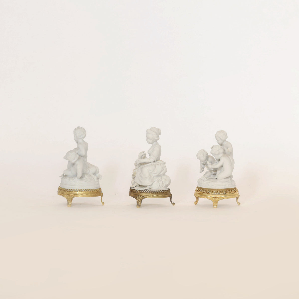 A Set of Porcelain Cherub Figurines on Ormolu Bases by Euro Trash Annie, featuring three small white figurines with delicate human forms, is displayed against a plain, light background.