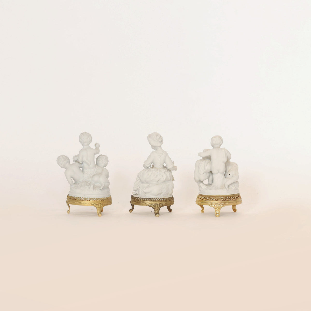 A set of Euro Trash Annie porcelain cherub figurines with ormolu bases is displayed in a row against a plain beige background, each piece featuring multiple figures in classical dress viewed from behind.
