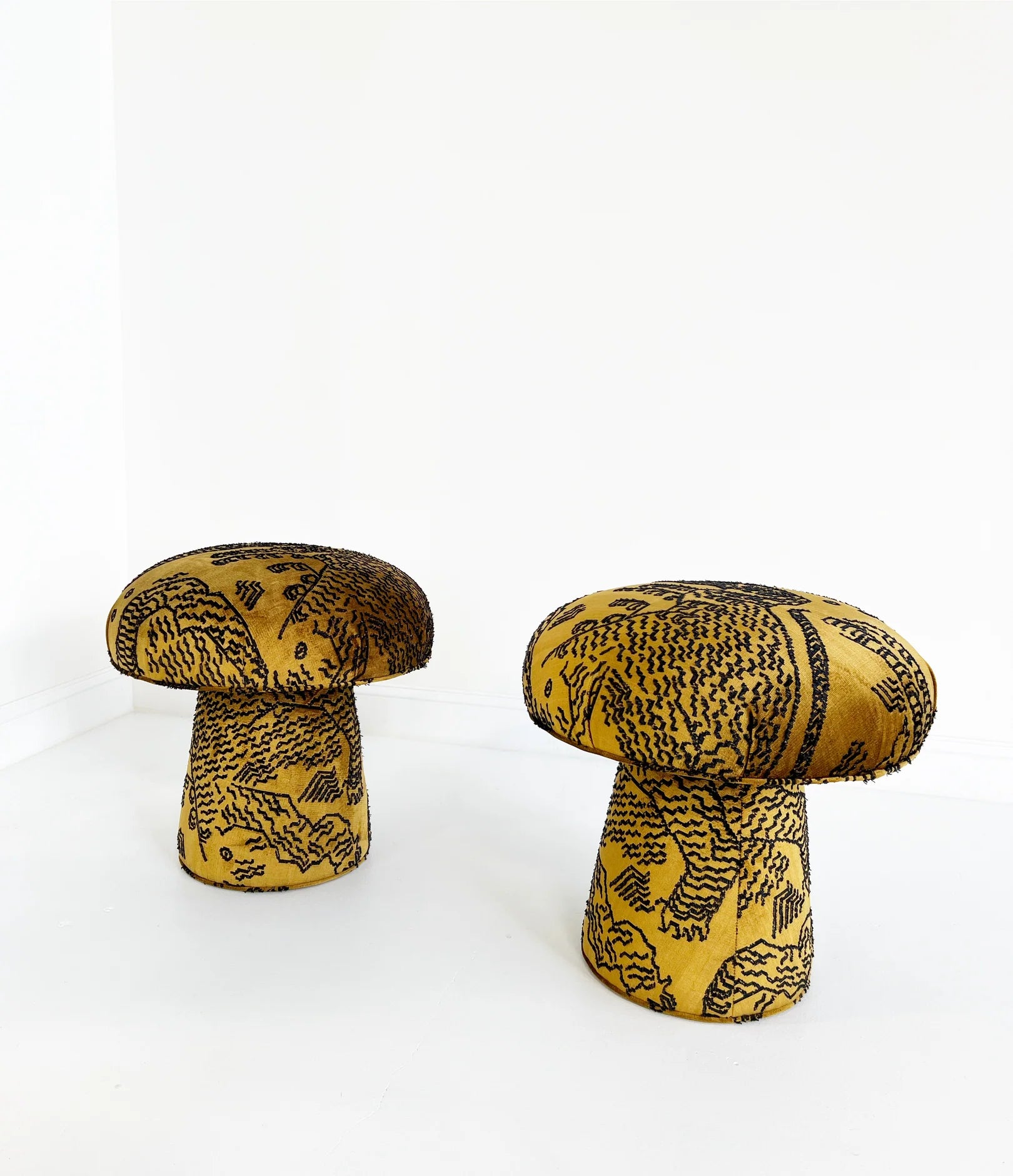 Two mushroom-shaped Forsyth Mushroom Poufs in Dedar 'Tiger Mountain' jacquard velvet, featuring gold with bold black animal patterns, stand out in a minimalistic white room with clean walls and floors.