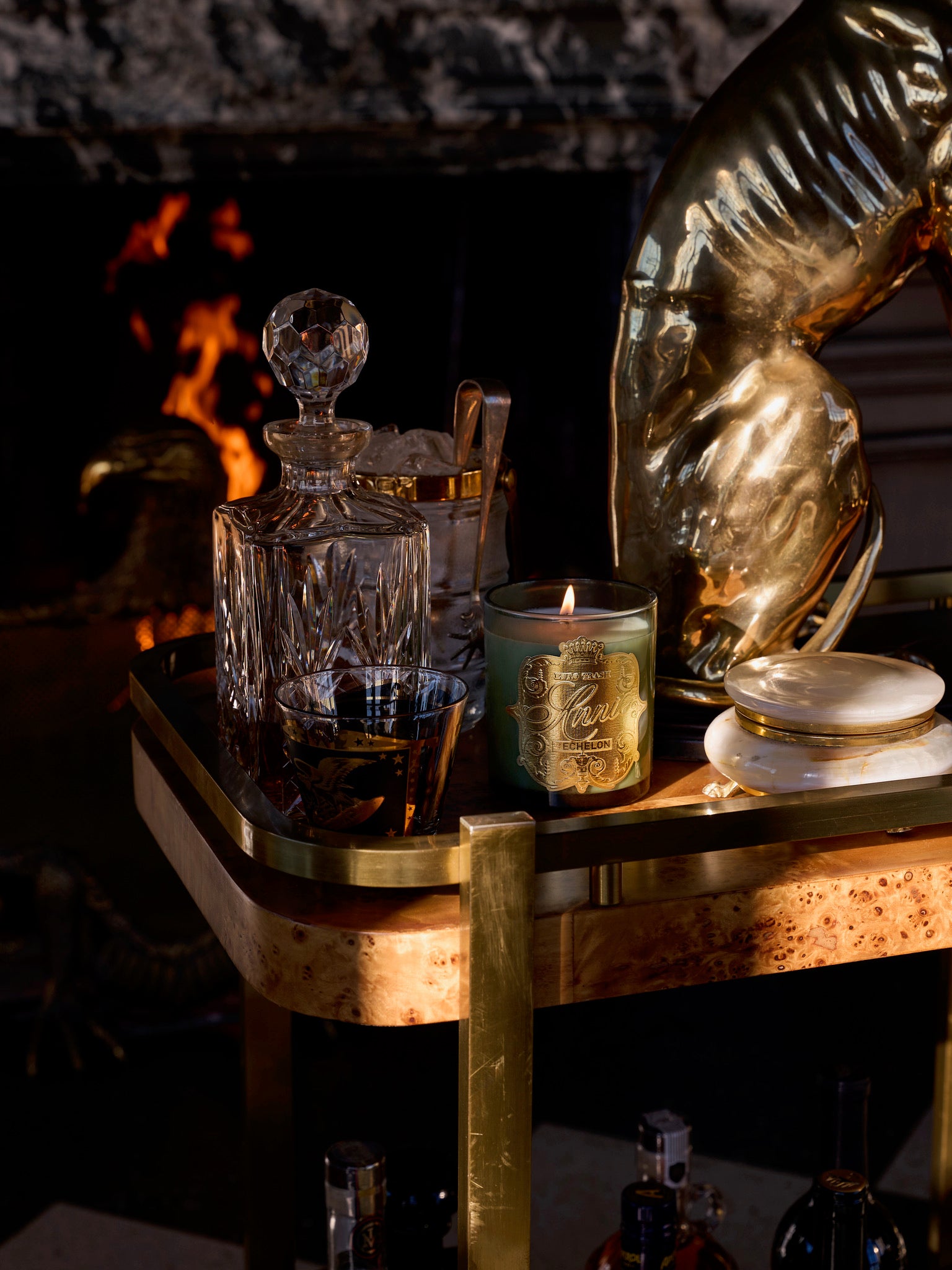 The Echelon, a crystal decanter filled with dark liquor featuring leather notes, sits alongside a green candle and gold accents on a chic bar cart before a glowing fireplace, evoking warmth and elegance.
