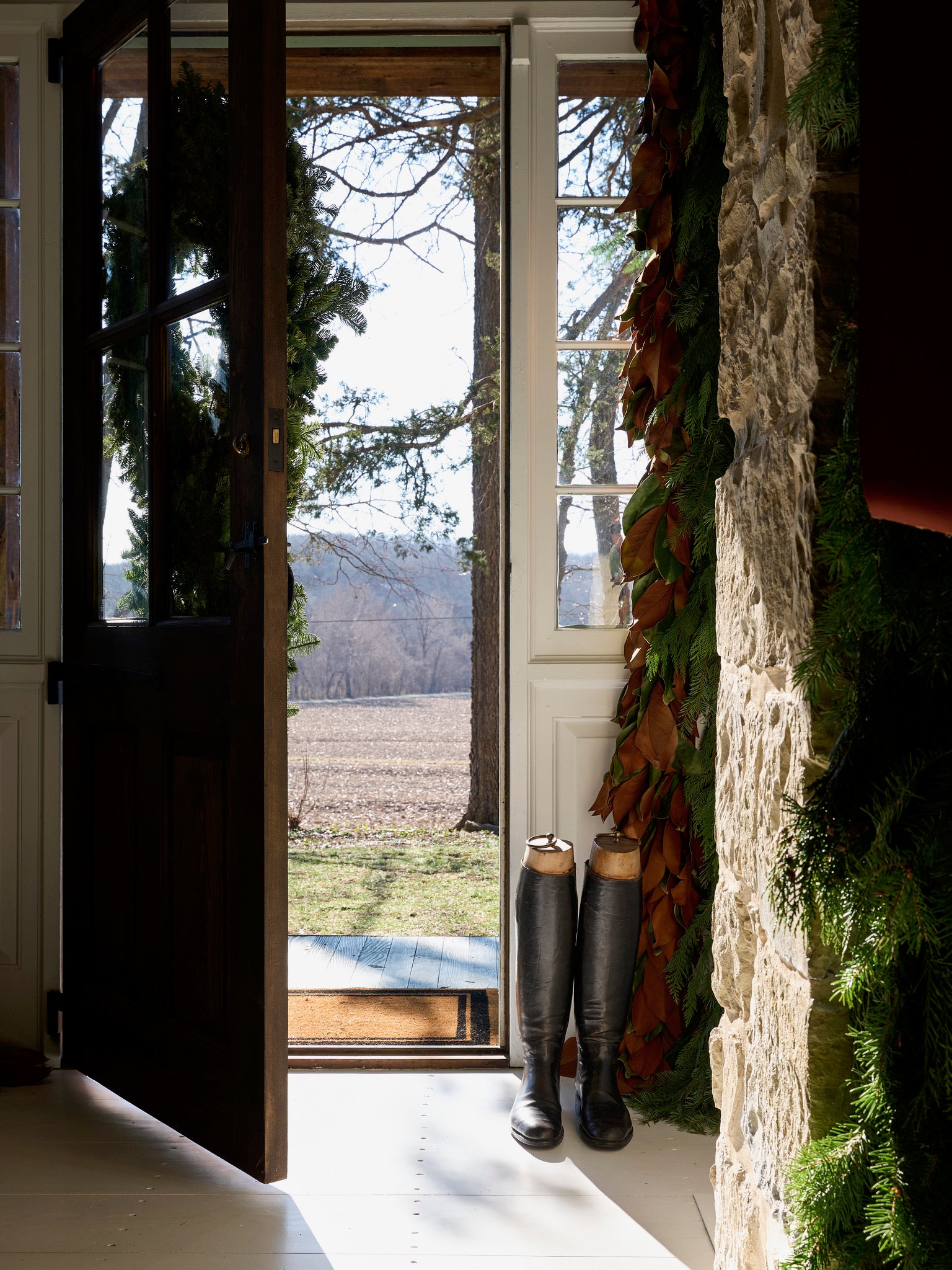 A wooden door is open to reveal a sunny outdoor scene with trees and a field. Black riding boots stand by the stone wall, which is decorated with green garland. Light streams into the cozy interior.