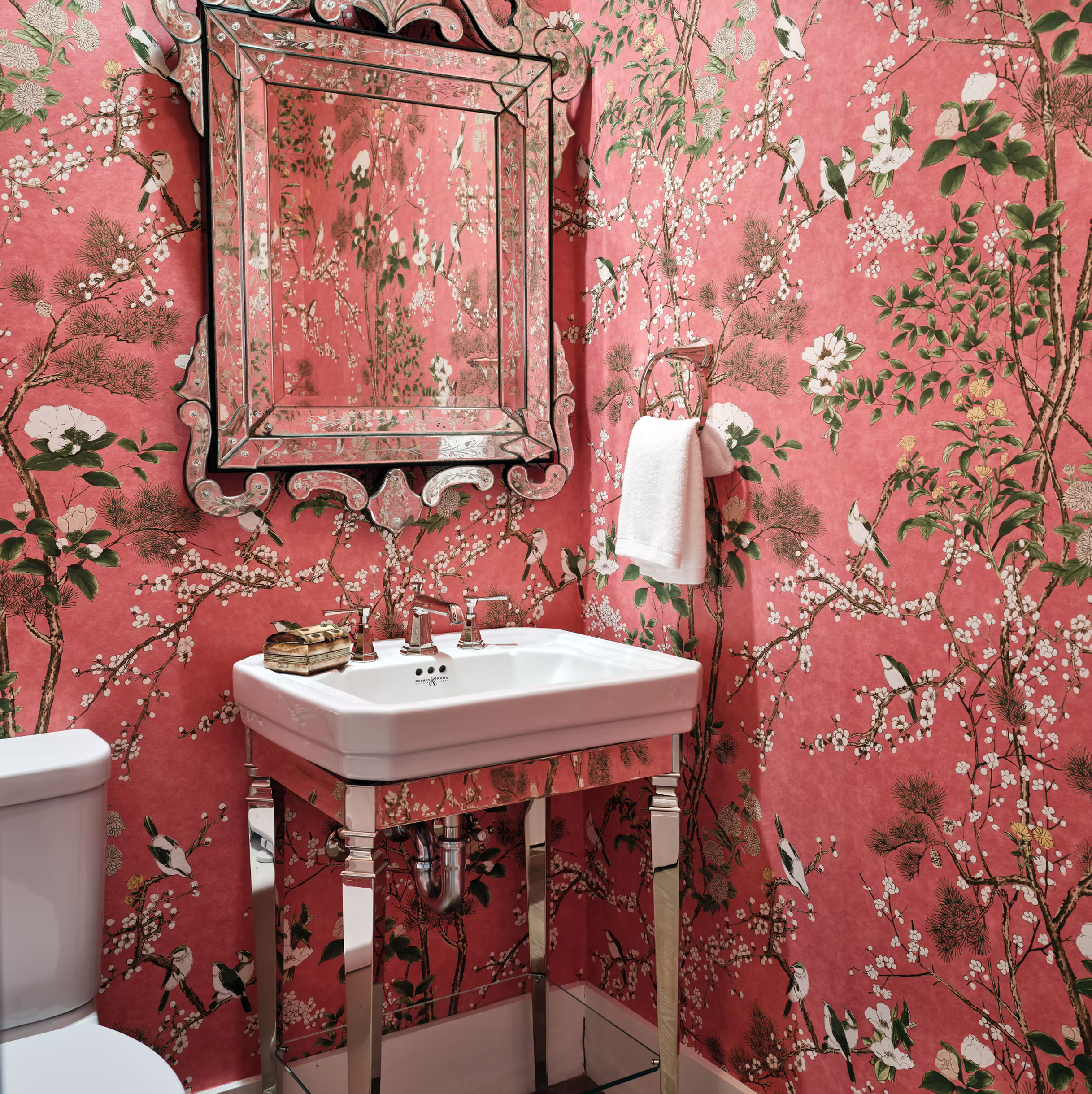 A small bathroom with pink floral wallpaper, a decorative mirror, a white sink with chrome legs, a wall-mounted towel ring with a white towel, and part of a toilet visible on the left.