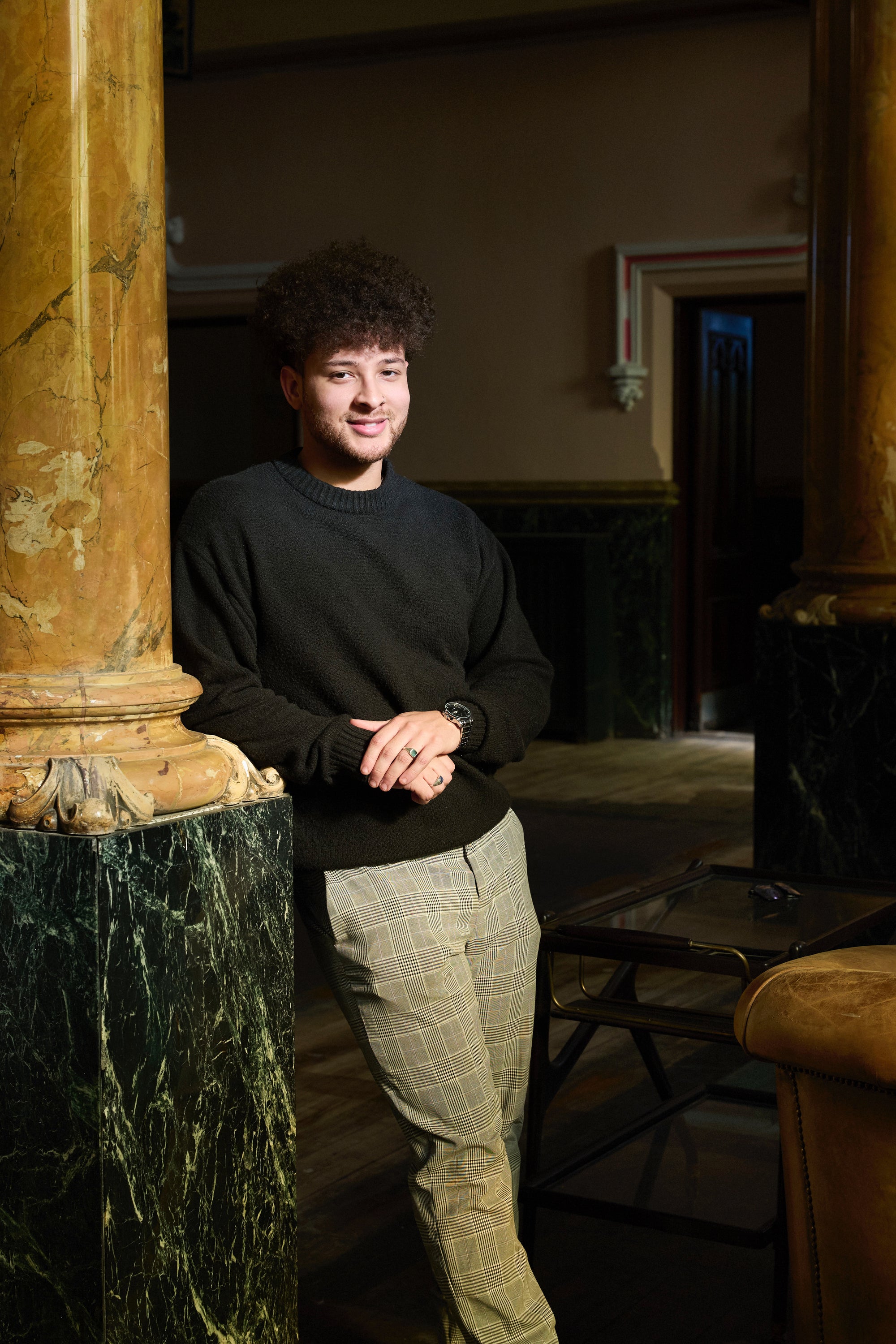 A man with curly hair and a beard, wearing a black sweater and plaid pants, leans against a marble column in an elegant, warmly lit room with dark wood furniture.