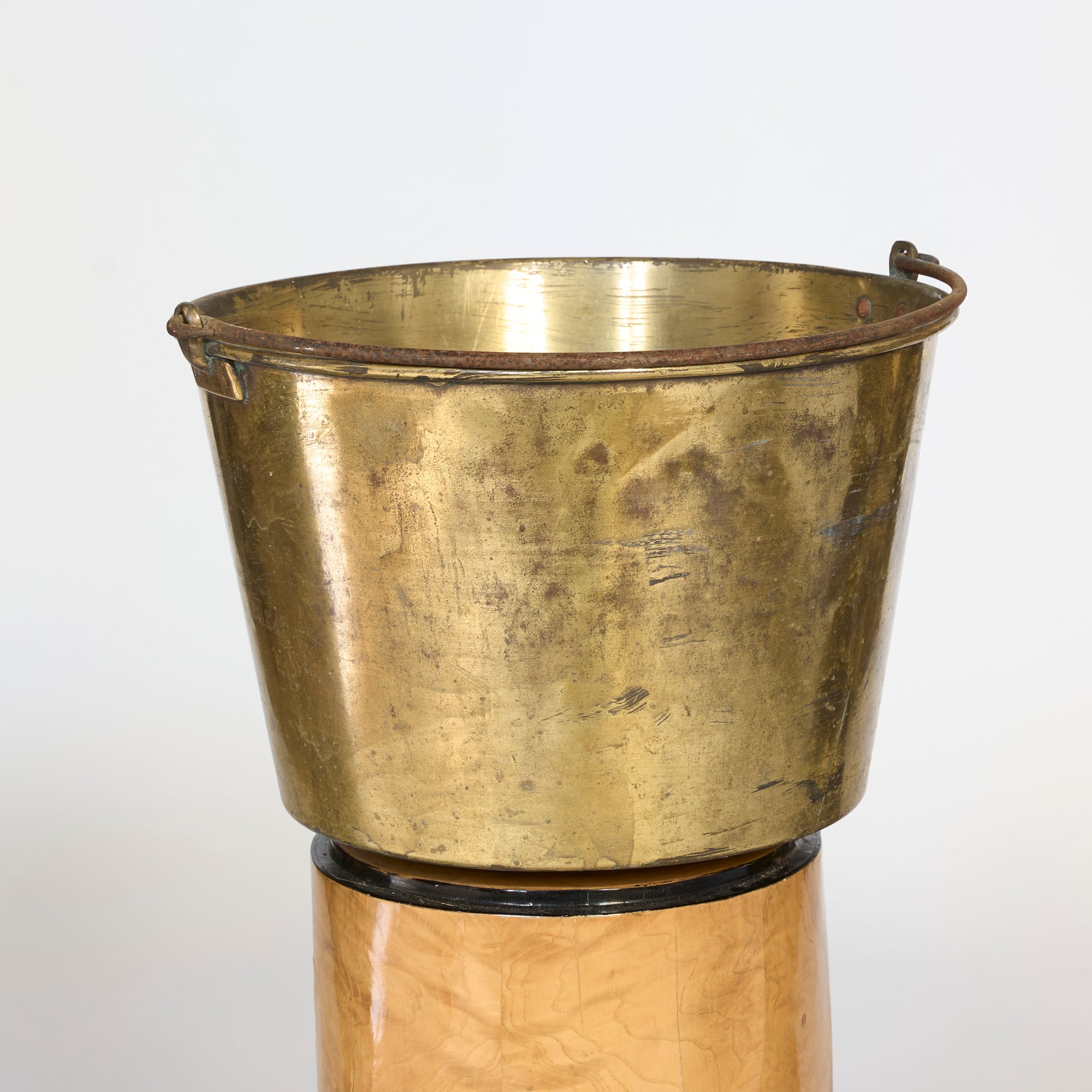 The Antique Brass Harvest Tub with Iron Handle sits atop a wooden cylindrical stand against a plain light background, showcasing charming tarnish and wear for vintage appeal.