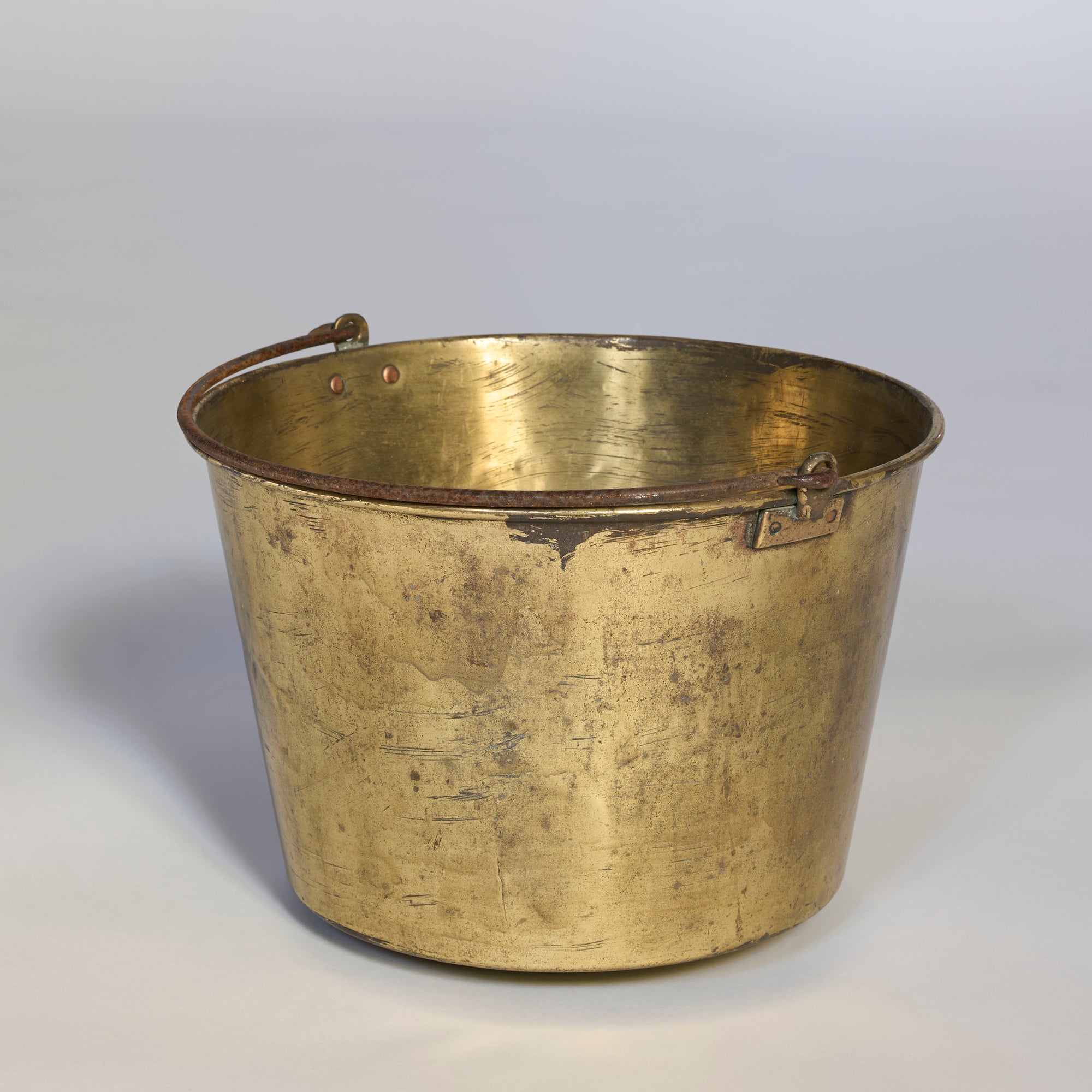 An Antique Brass Harvest Tub with an iron handle, featuring a shiny round design and visible scratches from use, sits on a smooth, neutral background.