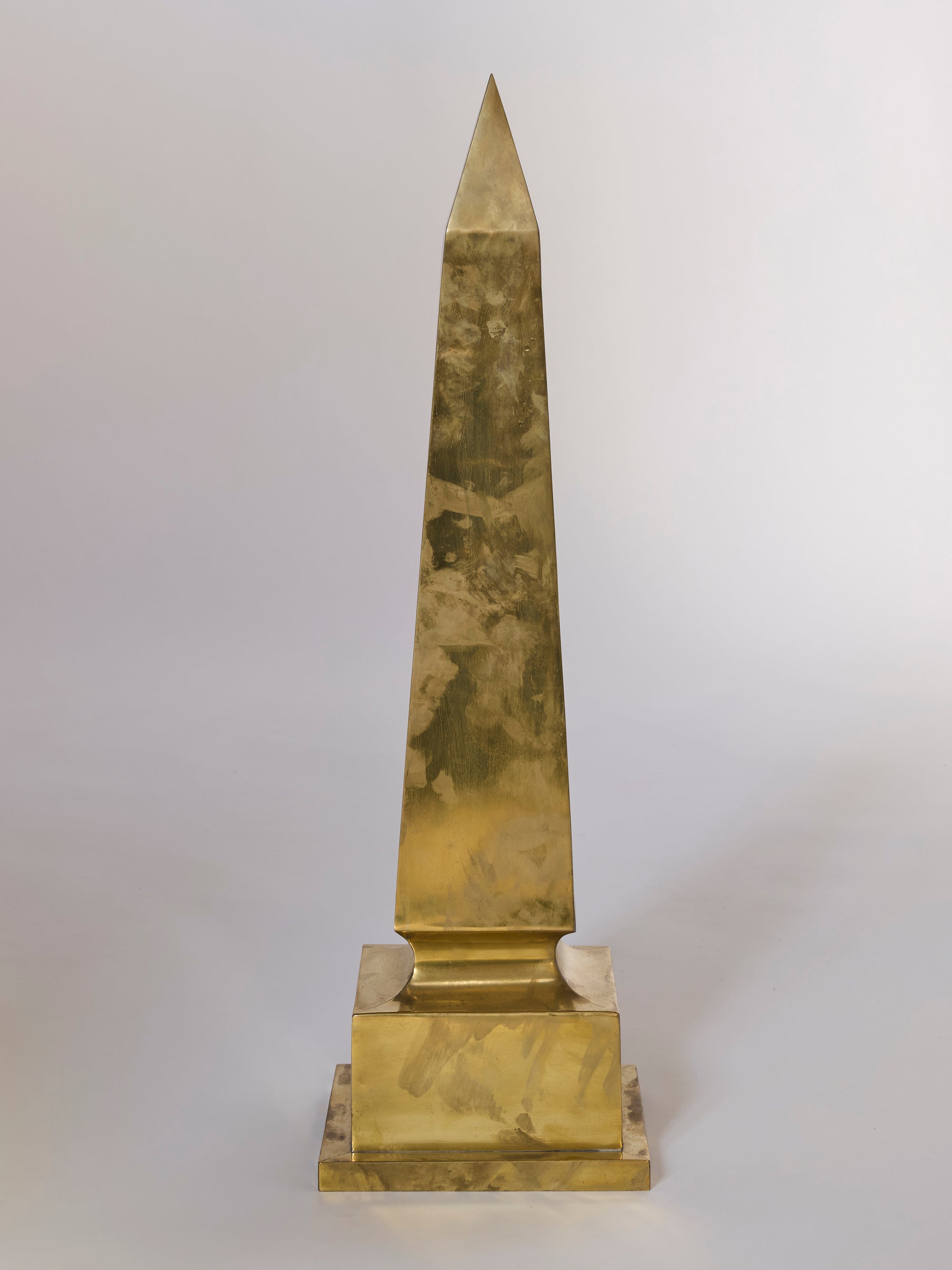 The Brass Obelisk Sculptural Objet d'art, with its square base and pointed top, stands out as a striking architectural statement piece against a plain light background in any modern decor.