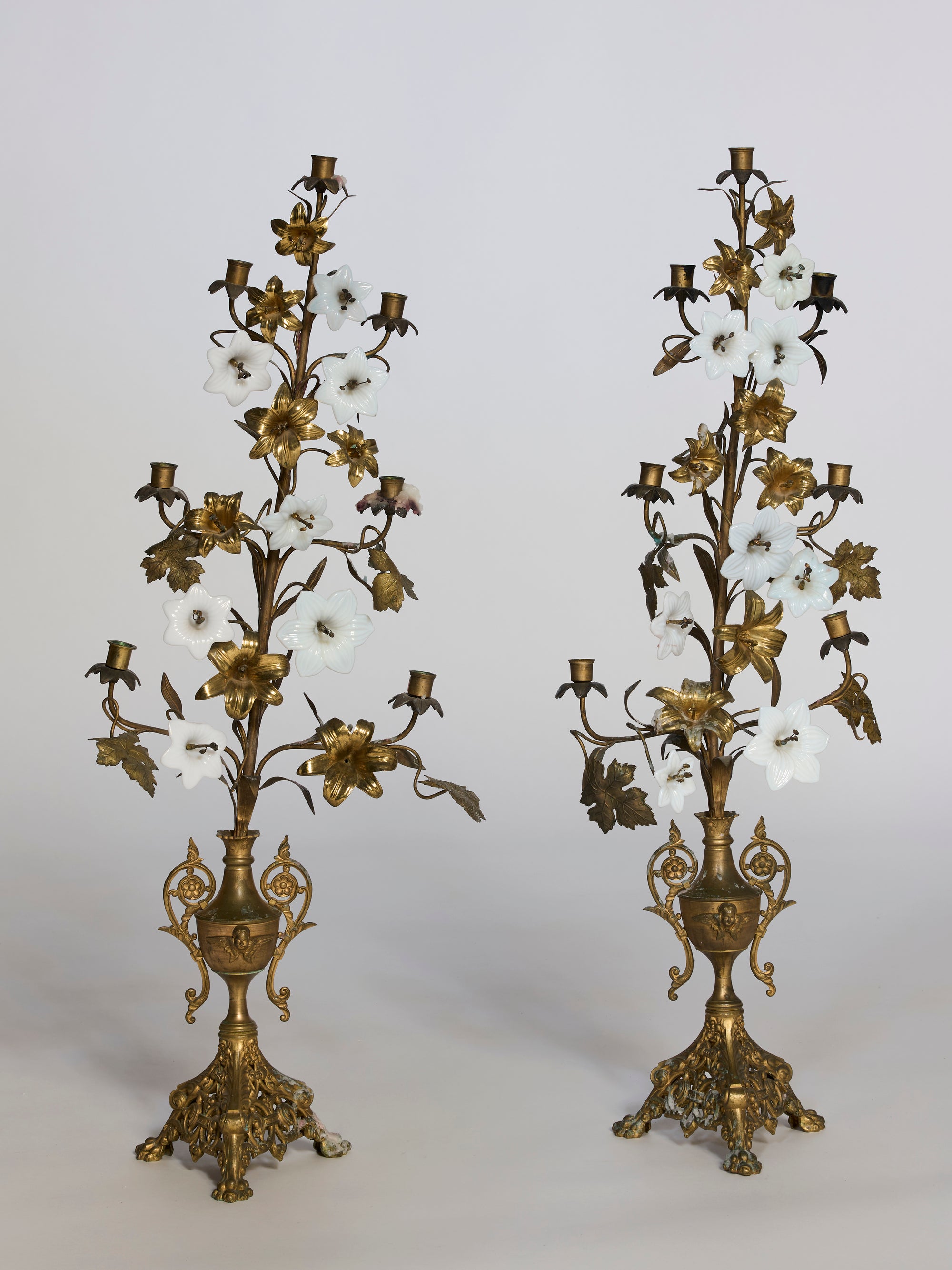 The Pair of Floral Brass Candelabra Sculptures feature ornate brass designs, white porcelain flowers, and multiple candle holders on elaborate pedestal bases—perfect for striking decorative display.