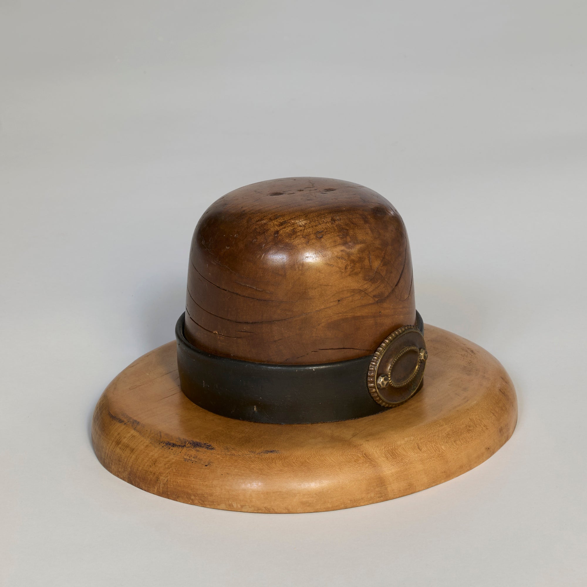 The Antique Wooden Millinery Hat Mold with Brass Detail, shaped like a bowler hat with a dark band and round brass buckle, sits on a flat wooden base against a light gray background, making it an attractive decorative piece.