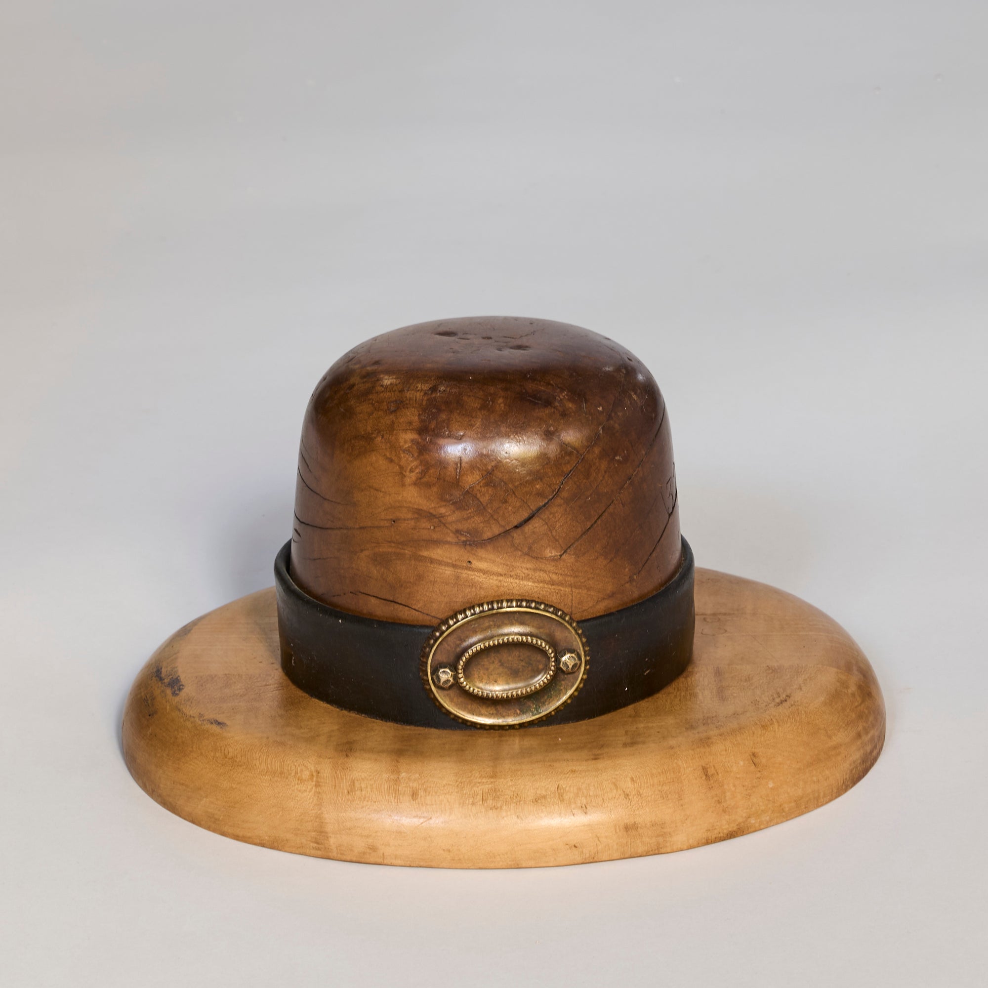 The Antique Wooden Millinery Hat Mold with Brass Detail is shaped like a wide-brimmed bowler hat with a black band and gold-tone buckle, making it an ideal decorative piece for collectors or millinery enthusiasts.