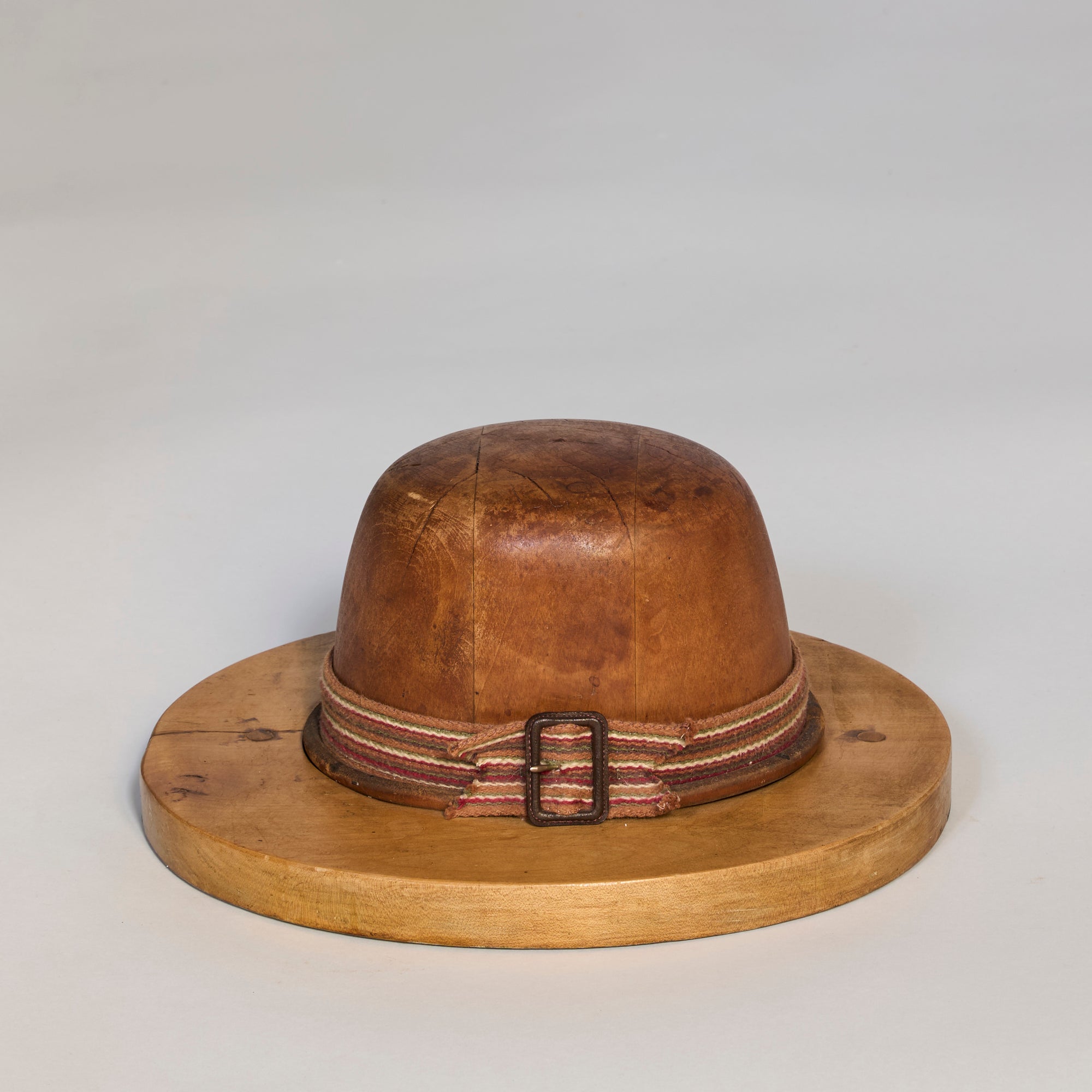 The Antique Wooden Hat Mold features a rounded crown and wide brim, accented by a striped fabric band with metal buckle, highlighting fine millinery craftsmanship against a light gray background.
