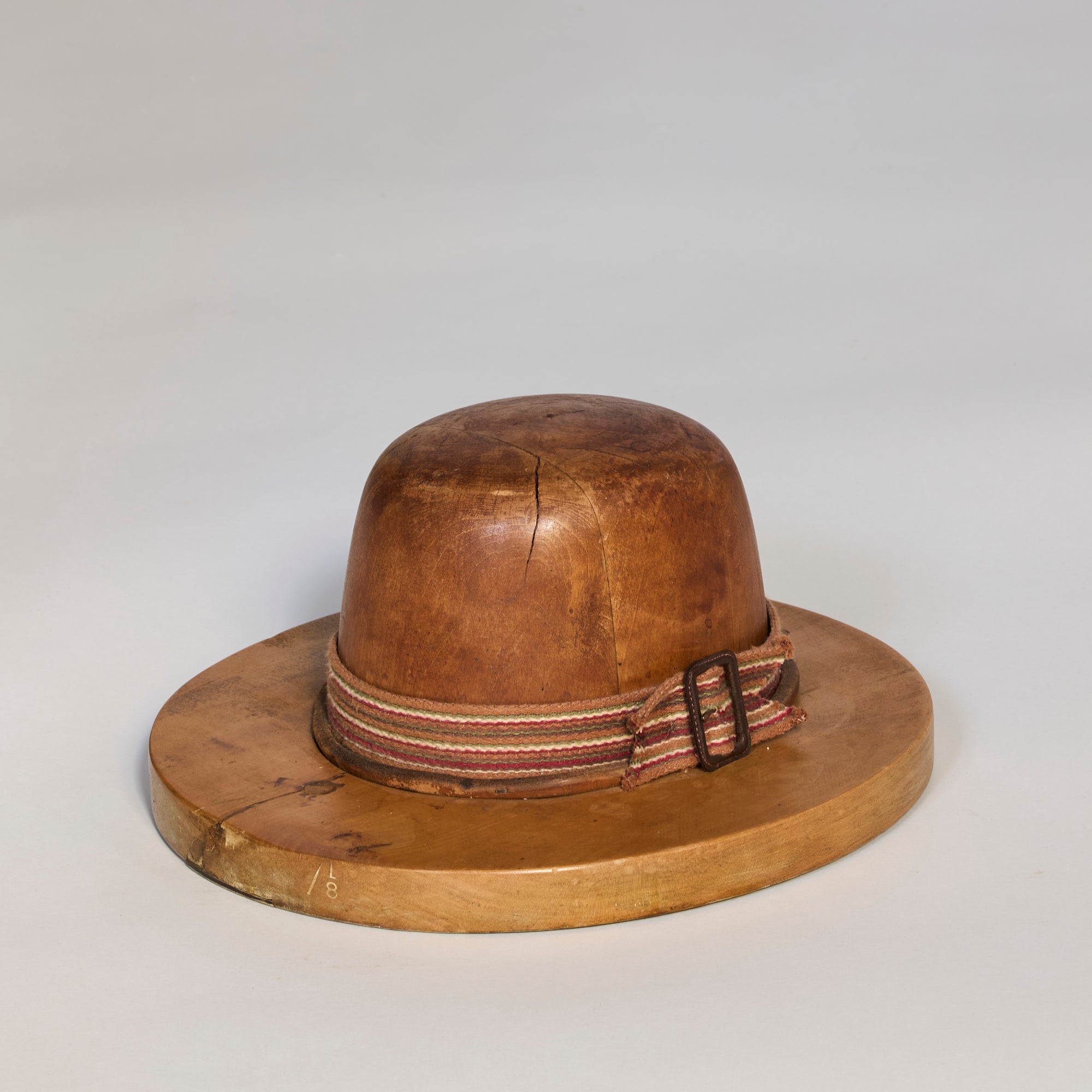 The Antique Wooden Hat Mold features a rounded crown and wide brim, displaying millinery craftsmanship with a striped fabric band and buckle at the crown's base, set against a plain light background.