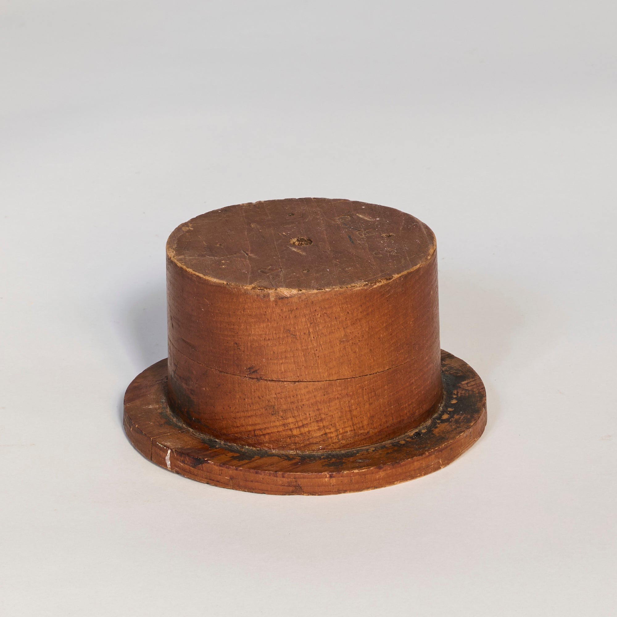 The Antique Wooden Hat Mold is cylindrical with a flat circular base, featuring worn wood, visible grain, and scratches. Shown on a plain light background, it makes an appealing decorative or sculptural interior piece.