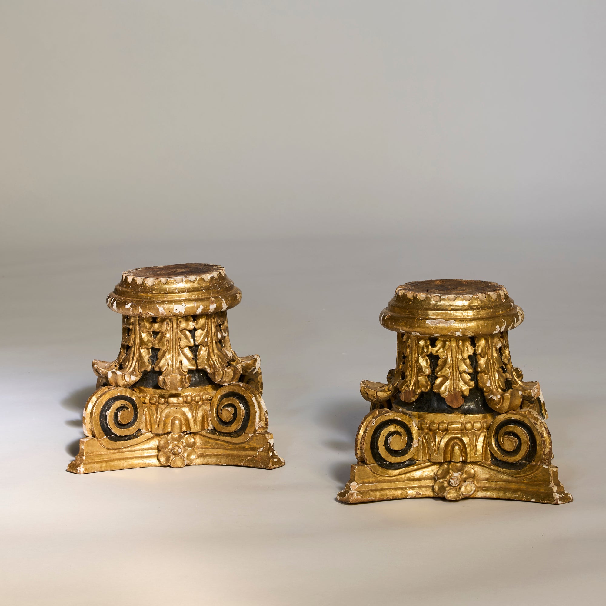 A Pair of Gilded Architectural Pedestal Bases featuring ornate woodwork, acanthus leaf carvings, and scrollwork stand against a plain, light background.
