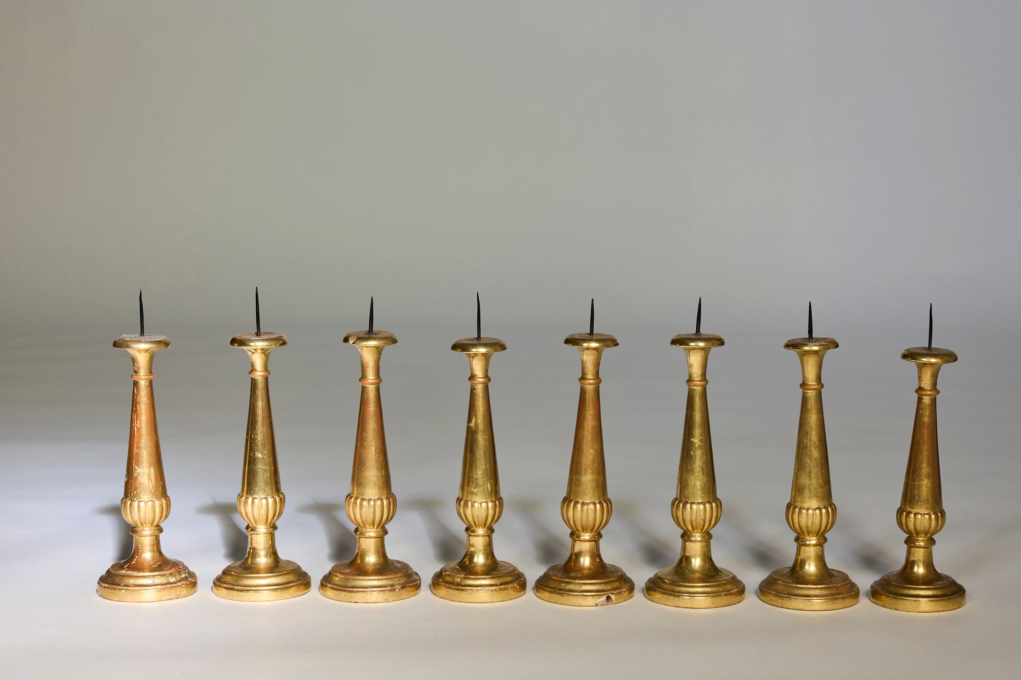 Eight Antique Gilt Candlearbra of varying heights and European designs are arranged in a row on a neutral background, each featuring a spike on top for holding candles.