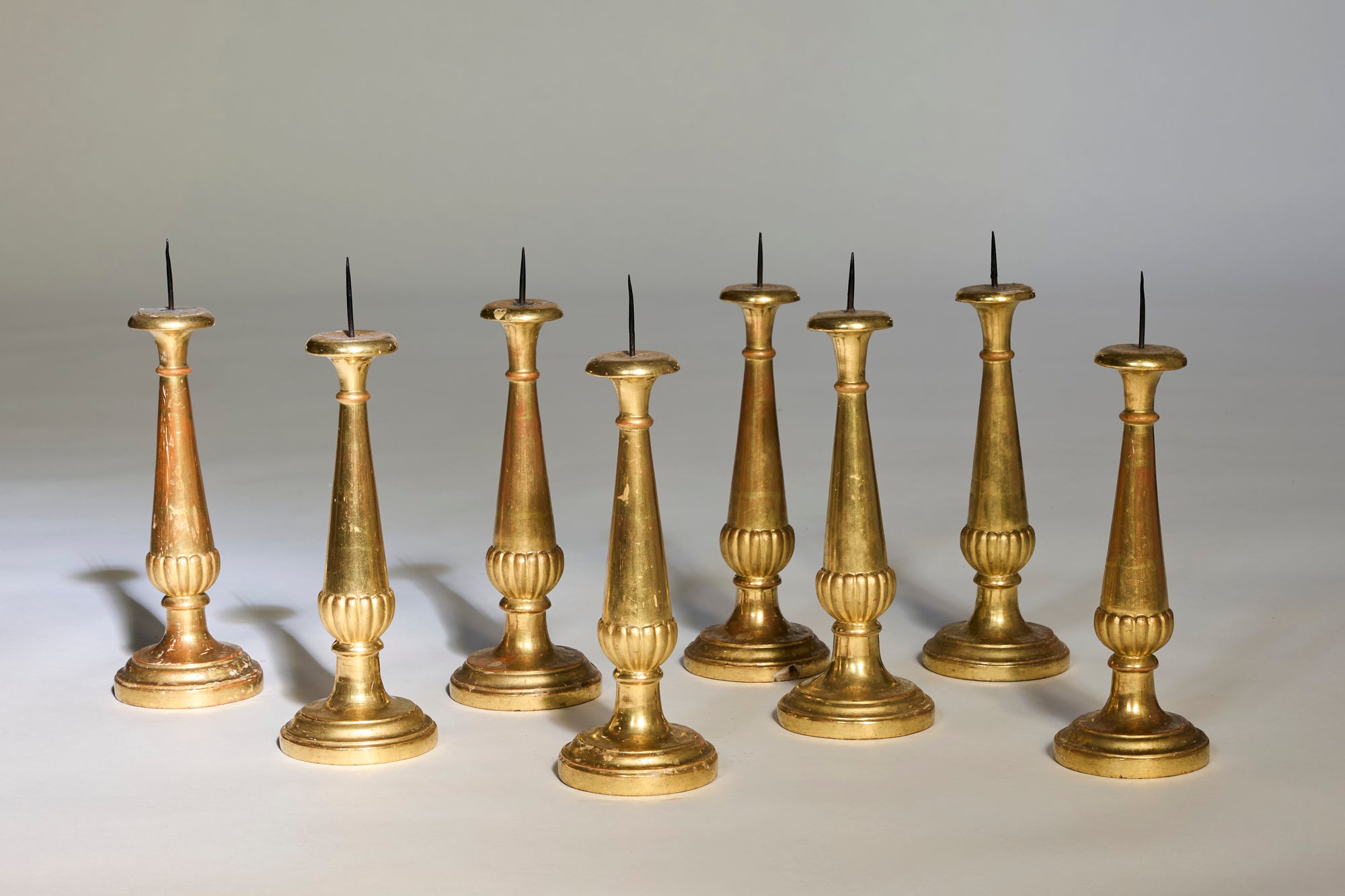 Eight Antique Gilt Candelabra, featuring tall, slender stems and round bases with sharp candle spikes, are arranged in two rows on a light surface—a fine example of European craftsmanship.