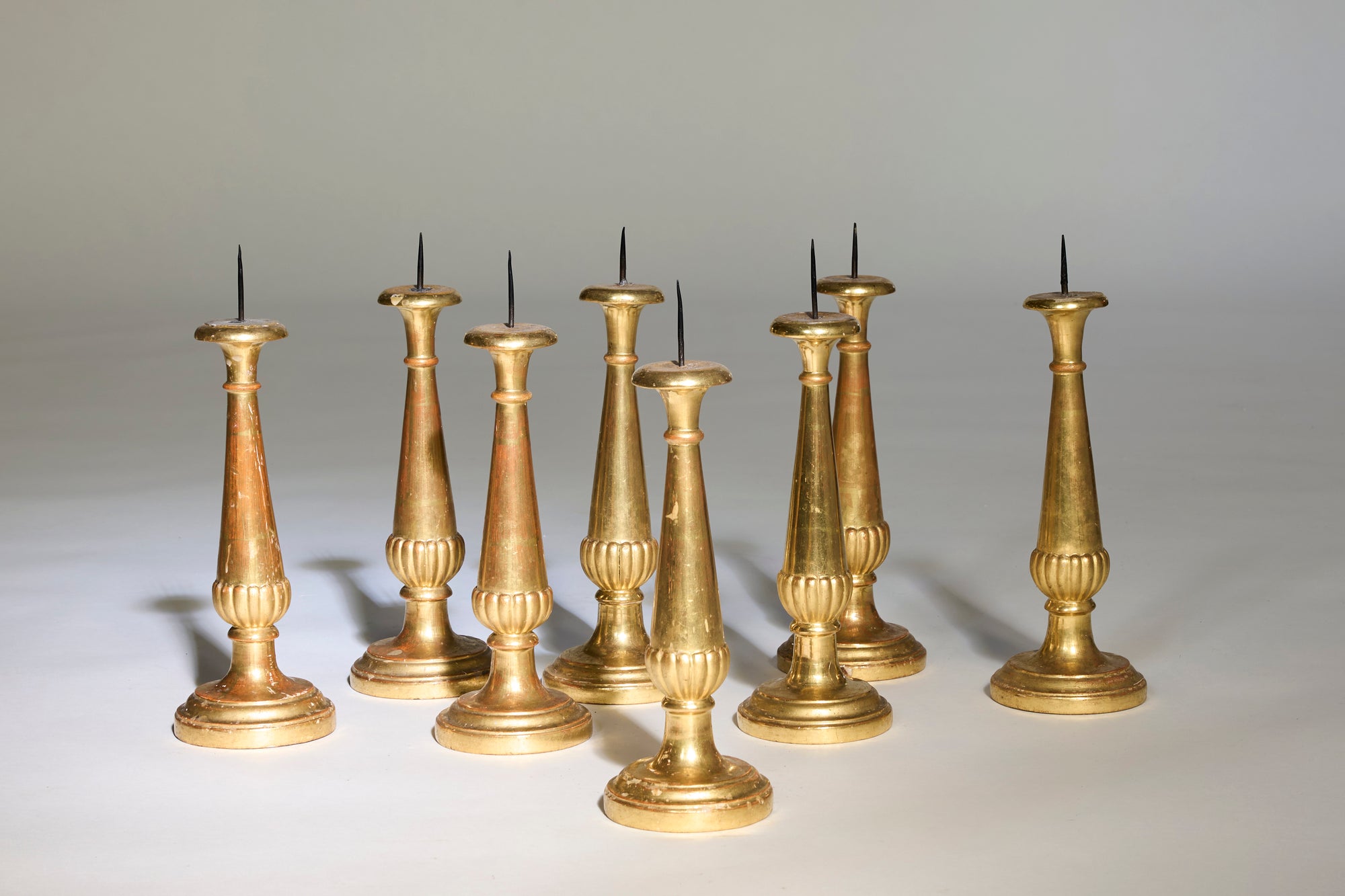 Eight Antique Gilt Candelabra with metal spikes, featuring ornate bases and varying heights, are displayed on a light surface. European craftsmanship shines in these elegant pieces set against a neutral background.