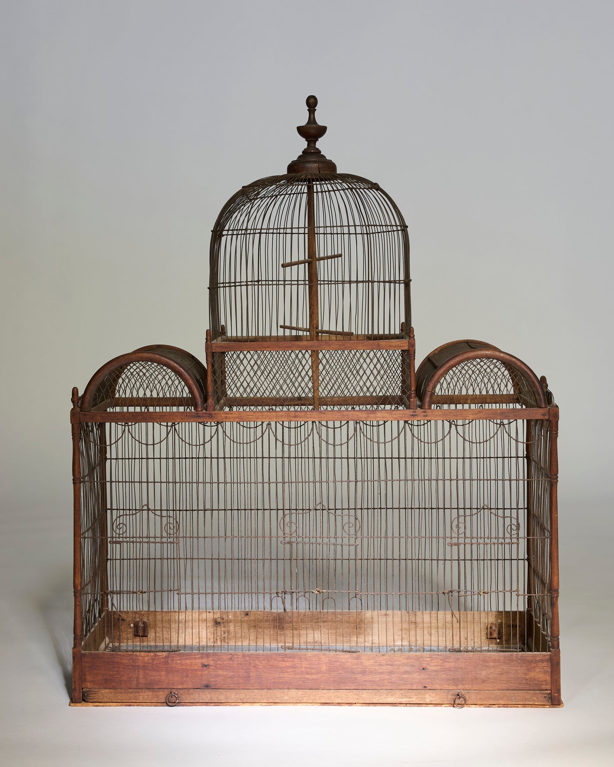 The Antique Architectural Wooden Birdcage Structure features a large brown metal frame with a domed top and twin arched sides, resting on a wooden base, creating a striking decorative piece against a plain background.