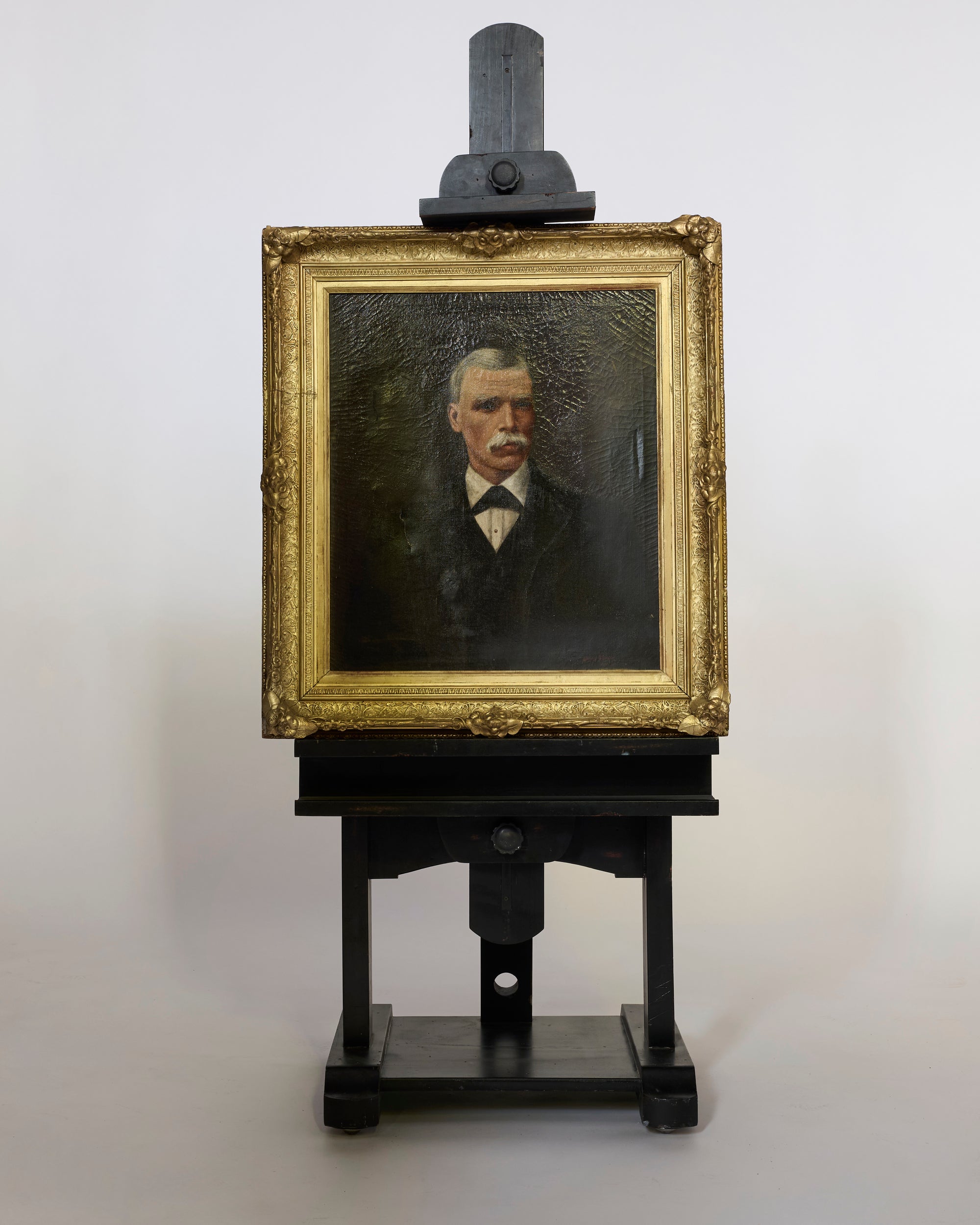 The Antique Portrait Oil Painting depicts a mustachioed man in a dark suit and bow tie, elegantly framed and displayed on a black wooden easel against a plain white background, radiating European refinement.