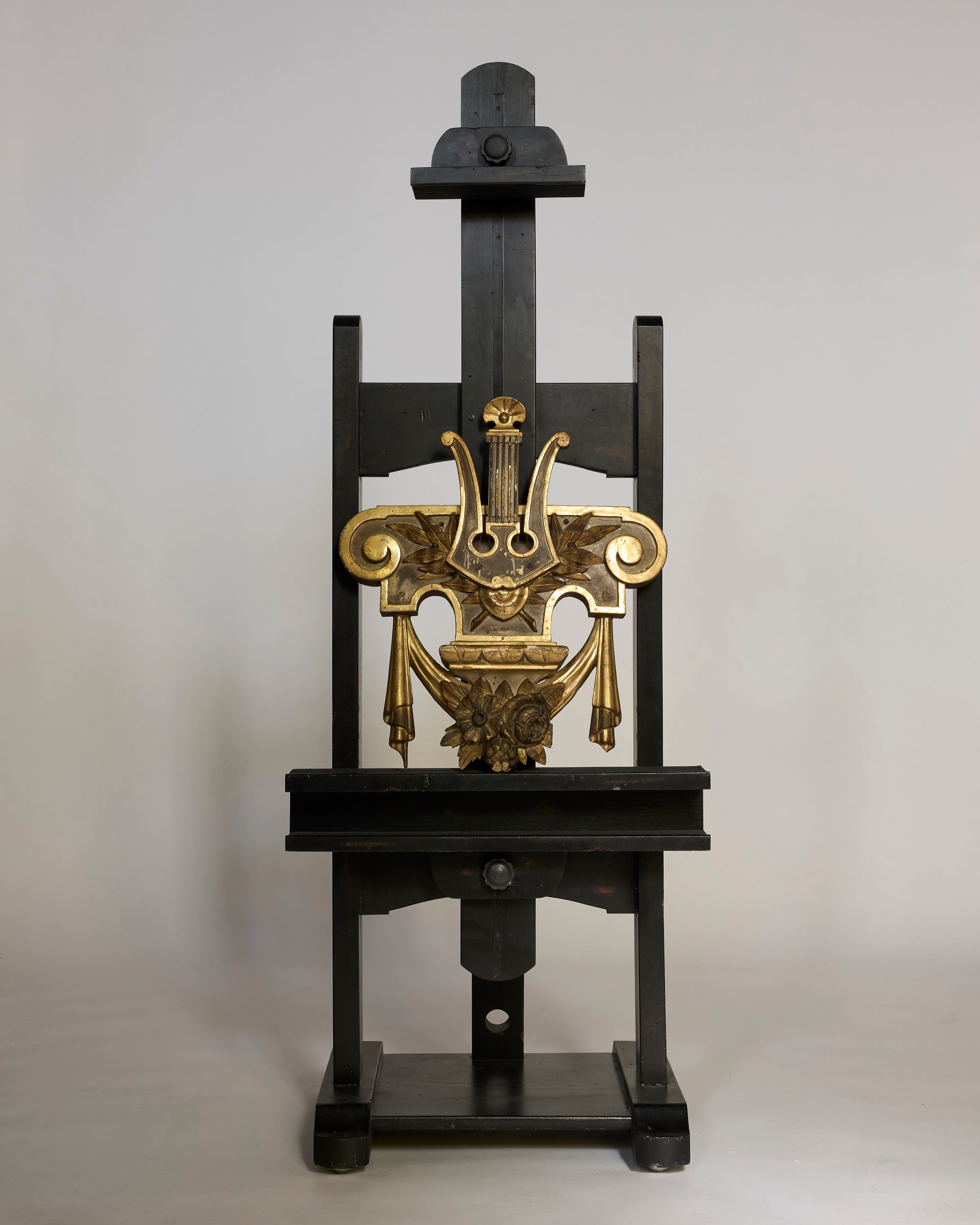 A Pair of Antique Giltwood Lyre & Foliate Architectural Ornaments is showcased on a tall black wooden easel against a plain, light gray background.