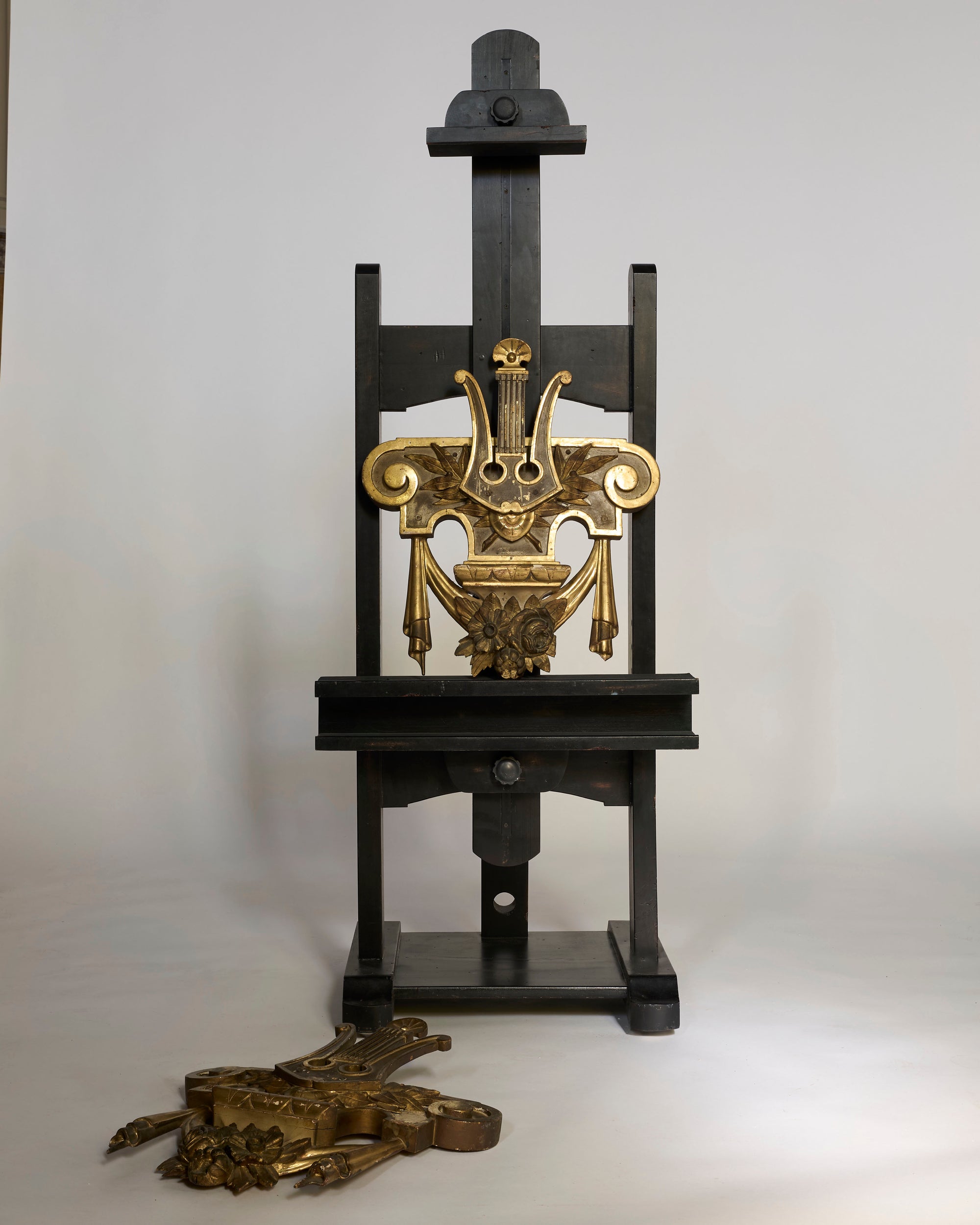 A black wooden easel displays a Pair of Antique Giltwood Lyre & Foliate Architectural Ornaments with intricate neoclassical details, while the second gilt ornament rests on the floor in front, set against a plain white background.