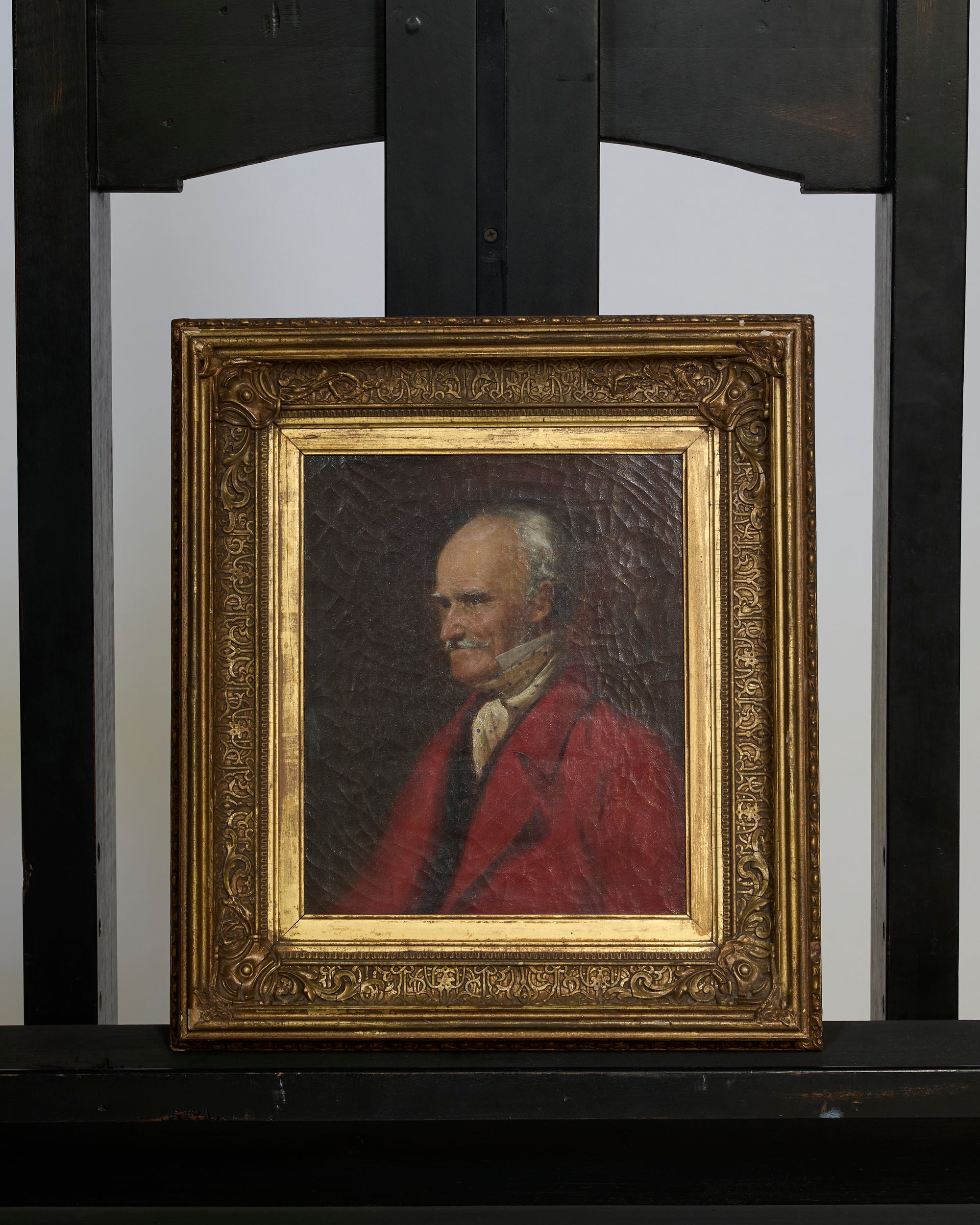 The Antique European Portrait Oil Painting depicts an older man in a red coat and white cravat set against a dark background, displayed on a black wooden chair with a decorative backrest.