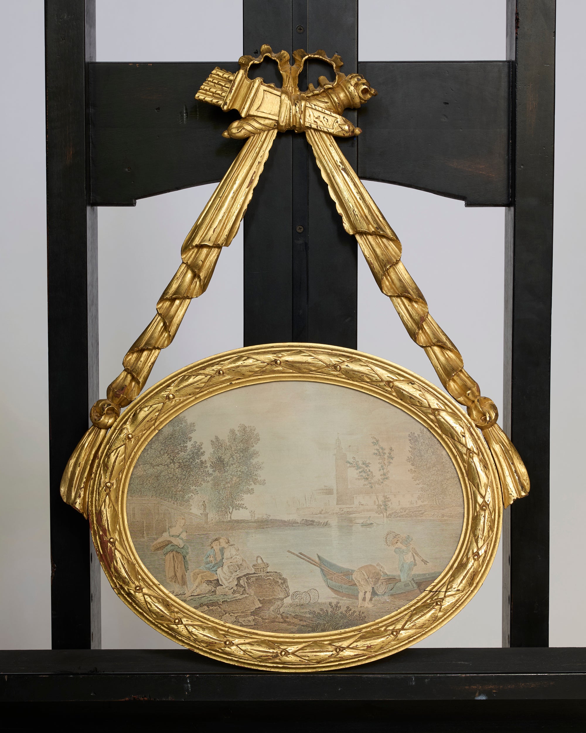 The Antique Giltwood Oval Pastoral Plaque with Carved Ribbon Swag features a hand-painted pastoral river scene with figures, buildings, and trees. The ornate plaque is displayed on a dark wooden easel.