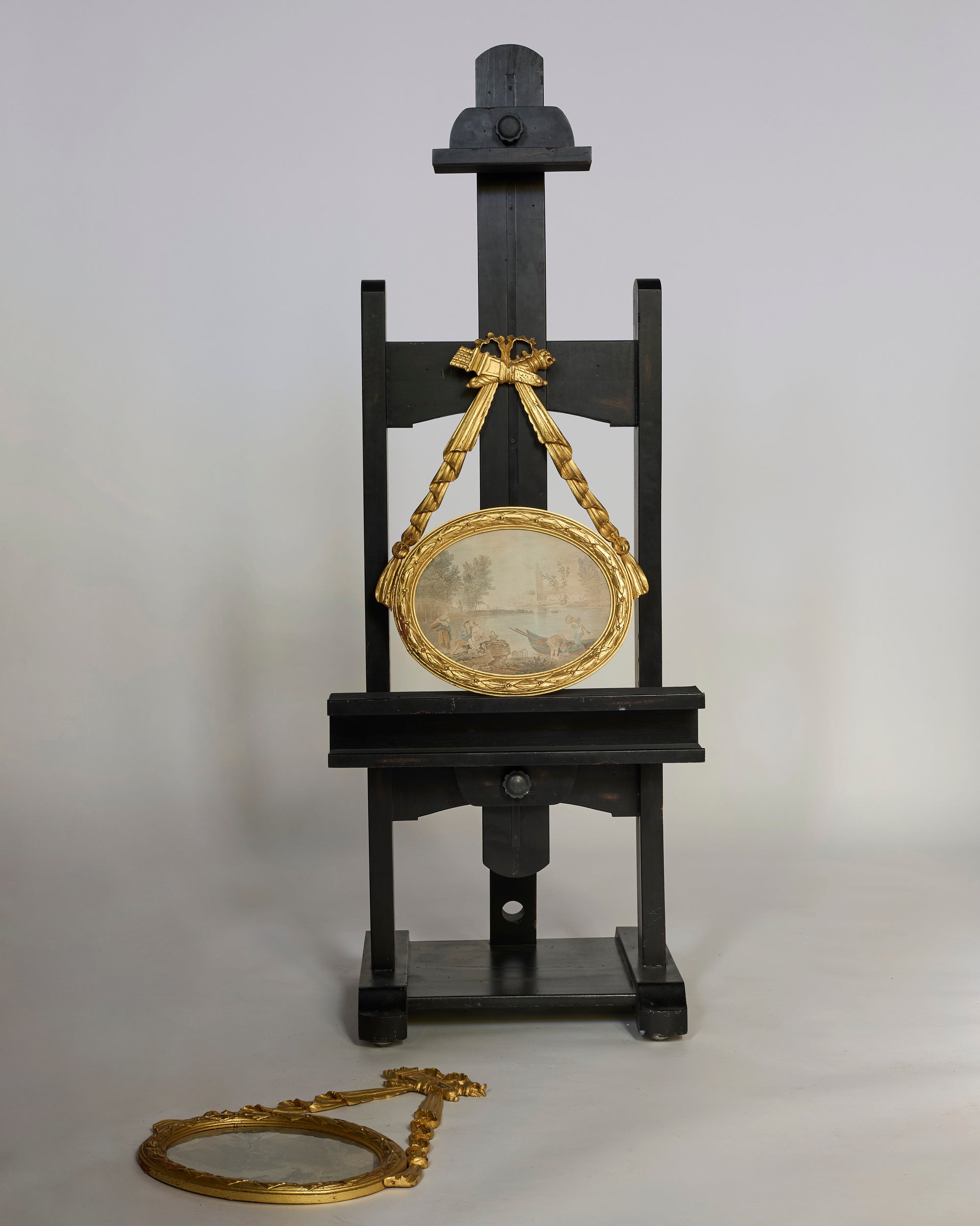 A black wooden easel displays an Antique Giltwood Oval Pastoral Plaque with Carved Ribbon Swag; a similar wall piece rests on the floor in front, both set against a simple light background.