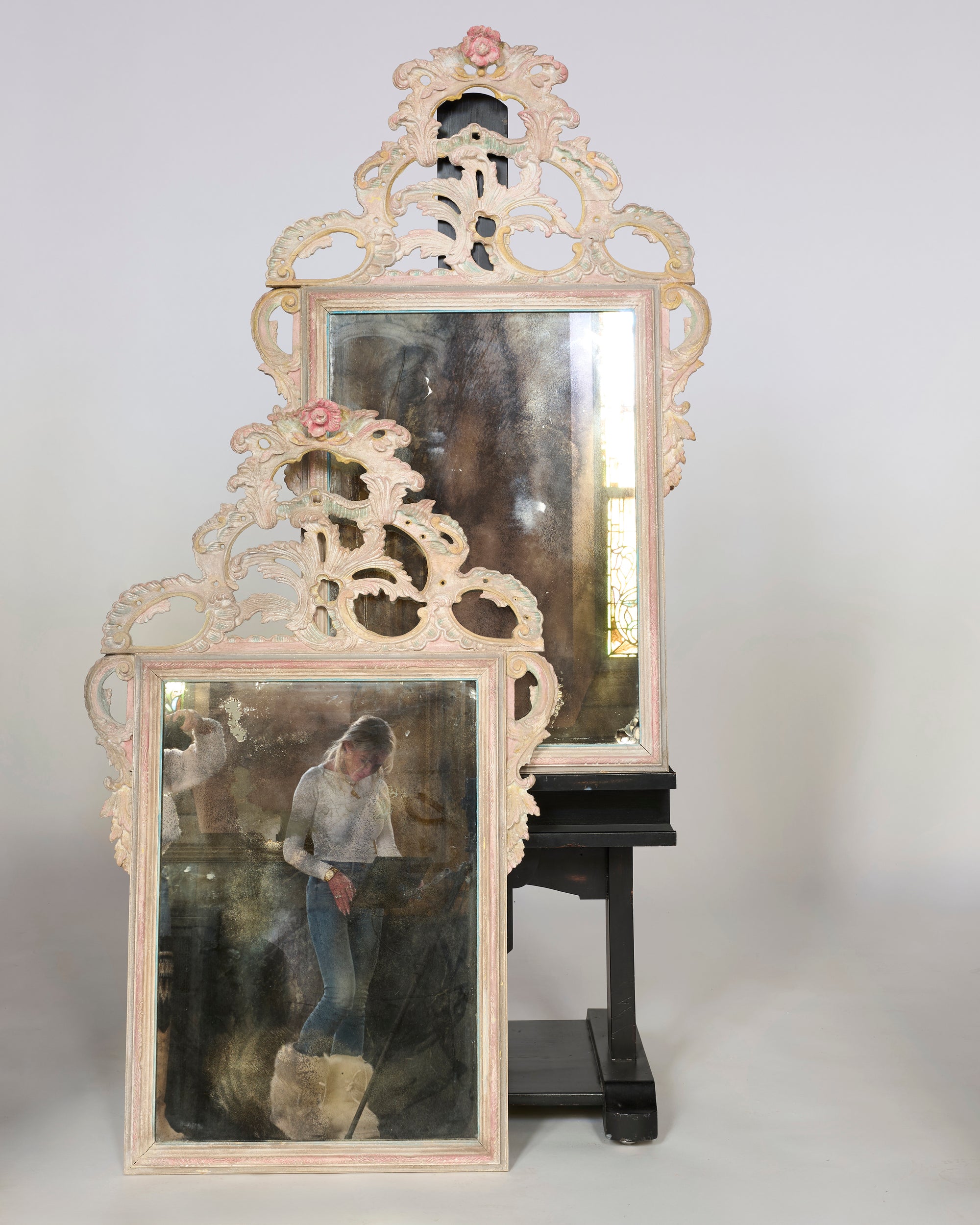 A Pair of Antique Rococo Style Carved Mirrors stand side by side; the lower mirror reflects a woman looking down, while the upper one shows a dark, indistinct room.