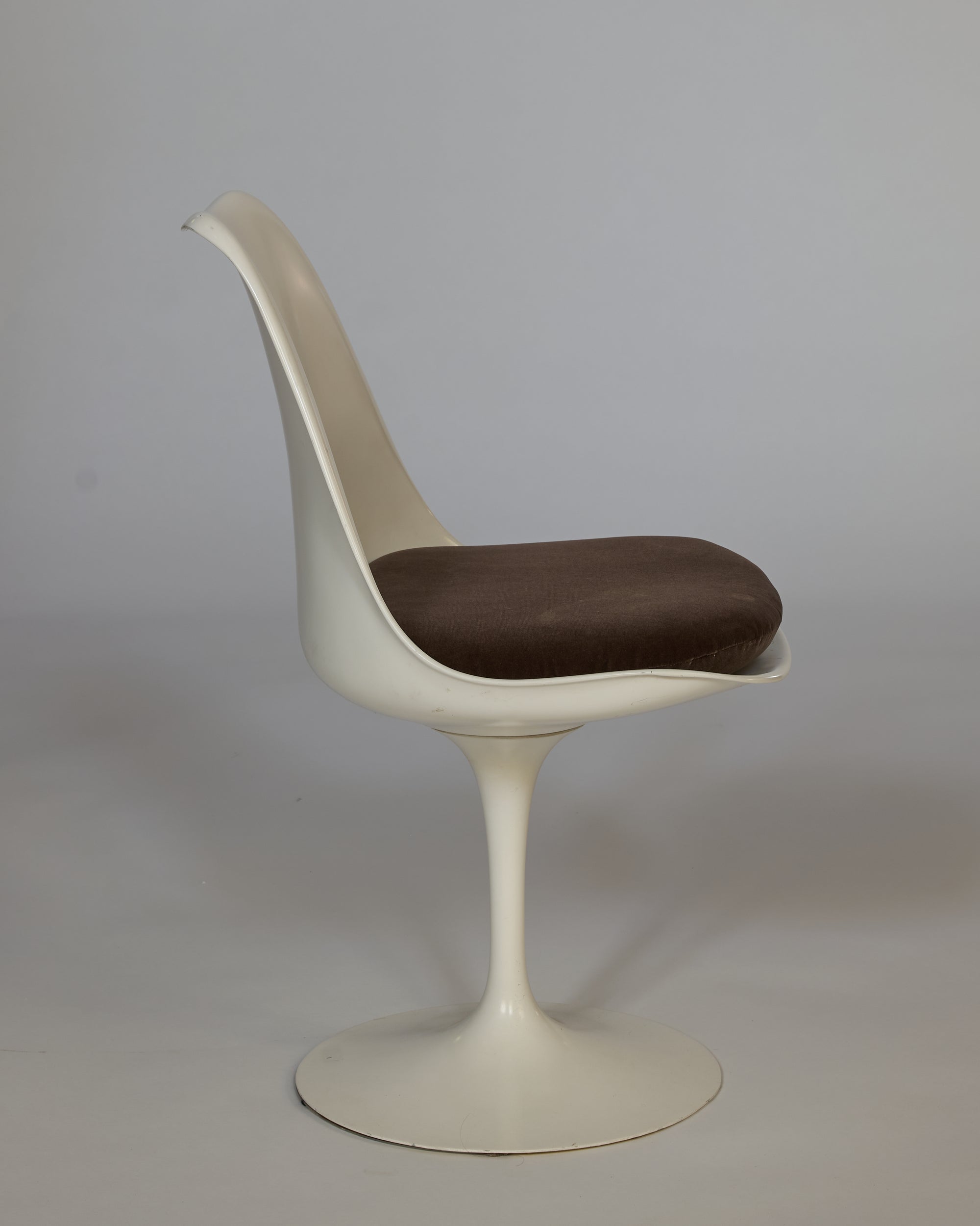The Vintage Mid-Century Modern Tulip Chair with Cushioned Seat is shown in side profile, featuring a white pedestal base, curved backrest, and round brown cushion against a plain gray background.