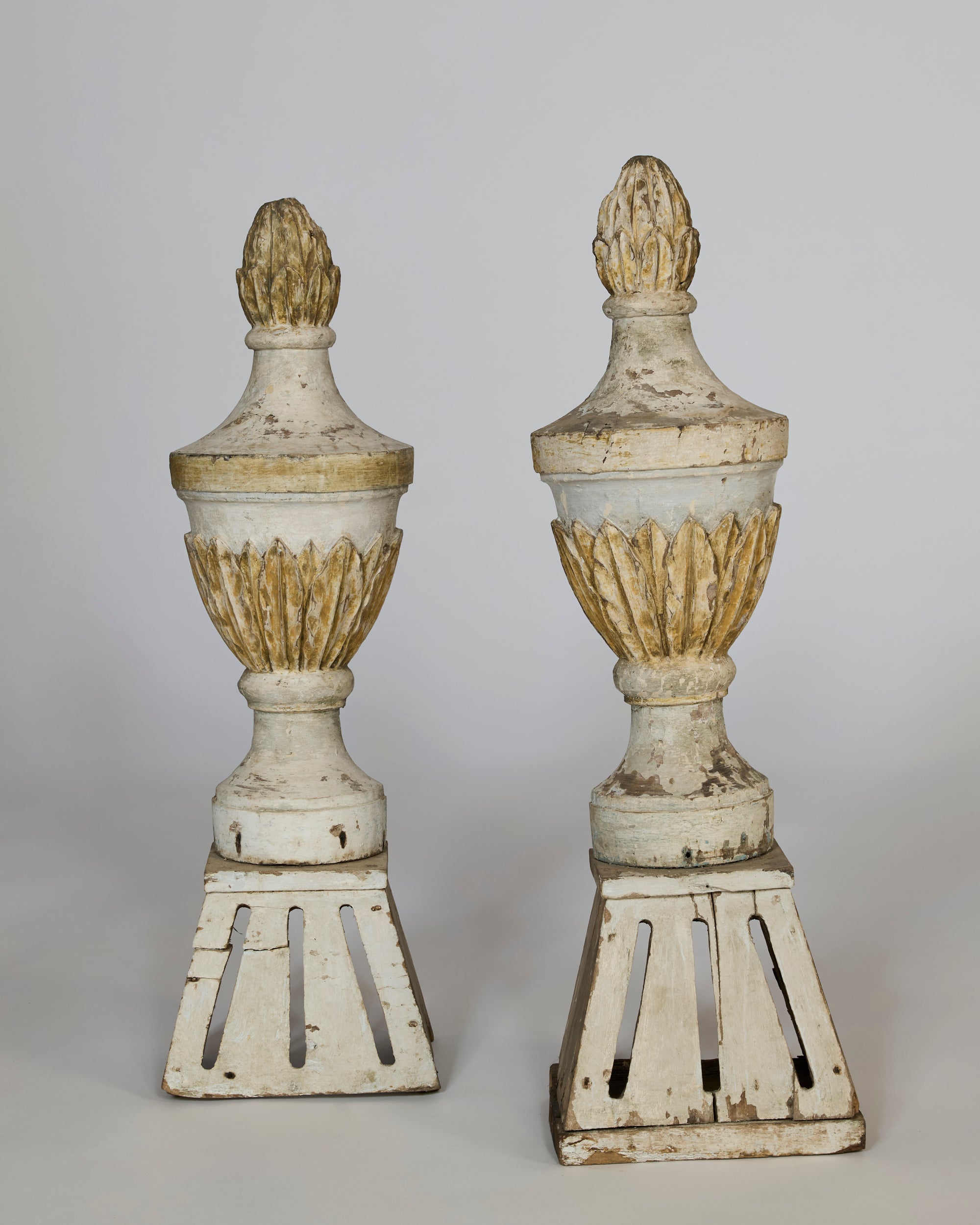 The Pair of Antique Carved Wooden Finials are weathered, hand-carved urn-shaped pieces with gold accents and flame-like tops, each standing on a square pedestal base against a plain background.
