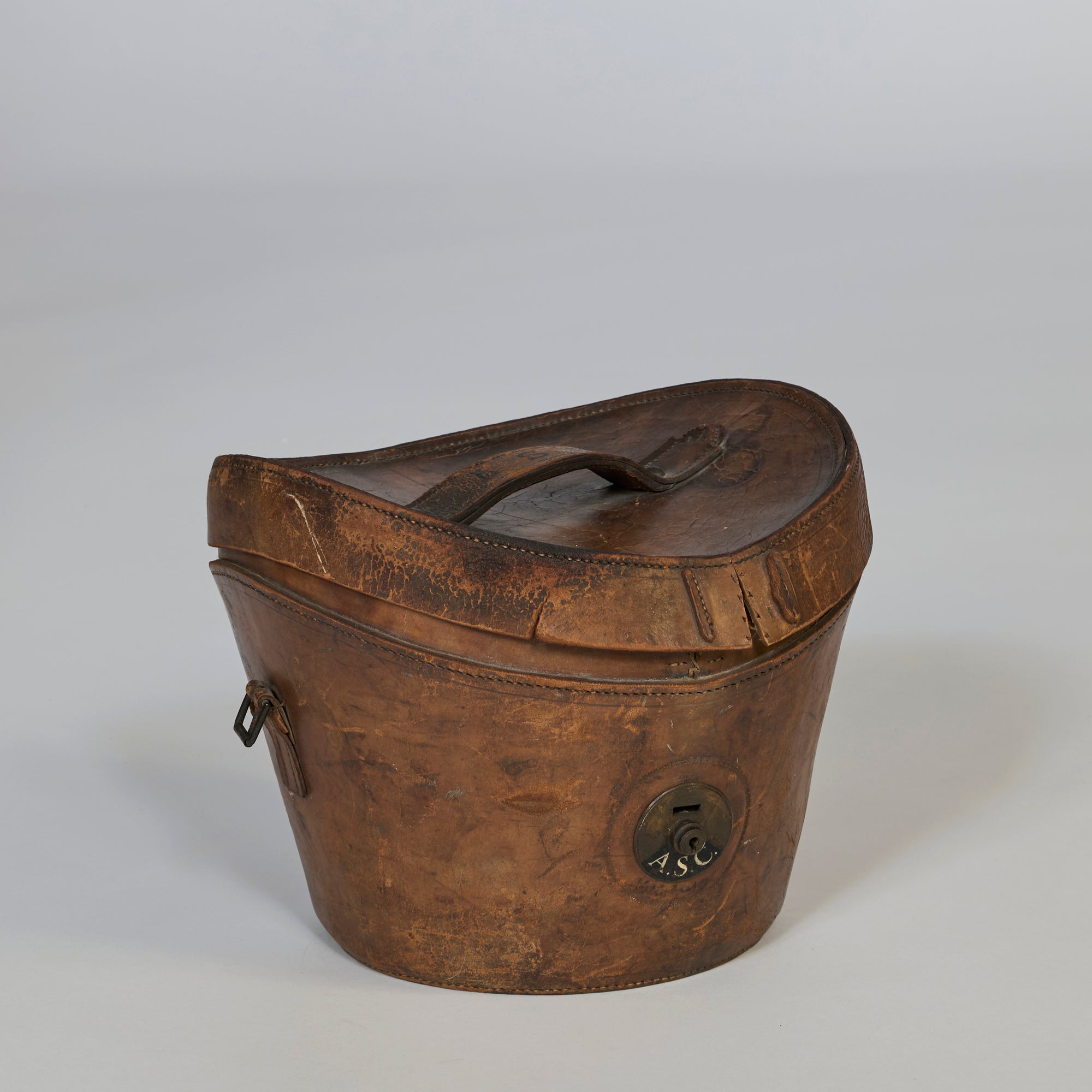 The Antique Leather Storage Case is a vintage brown leather hat box with a top handle, curved lid, side latch, and circular ASC-marked lock. It features heritage design and displays signs of age and wear.