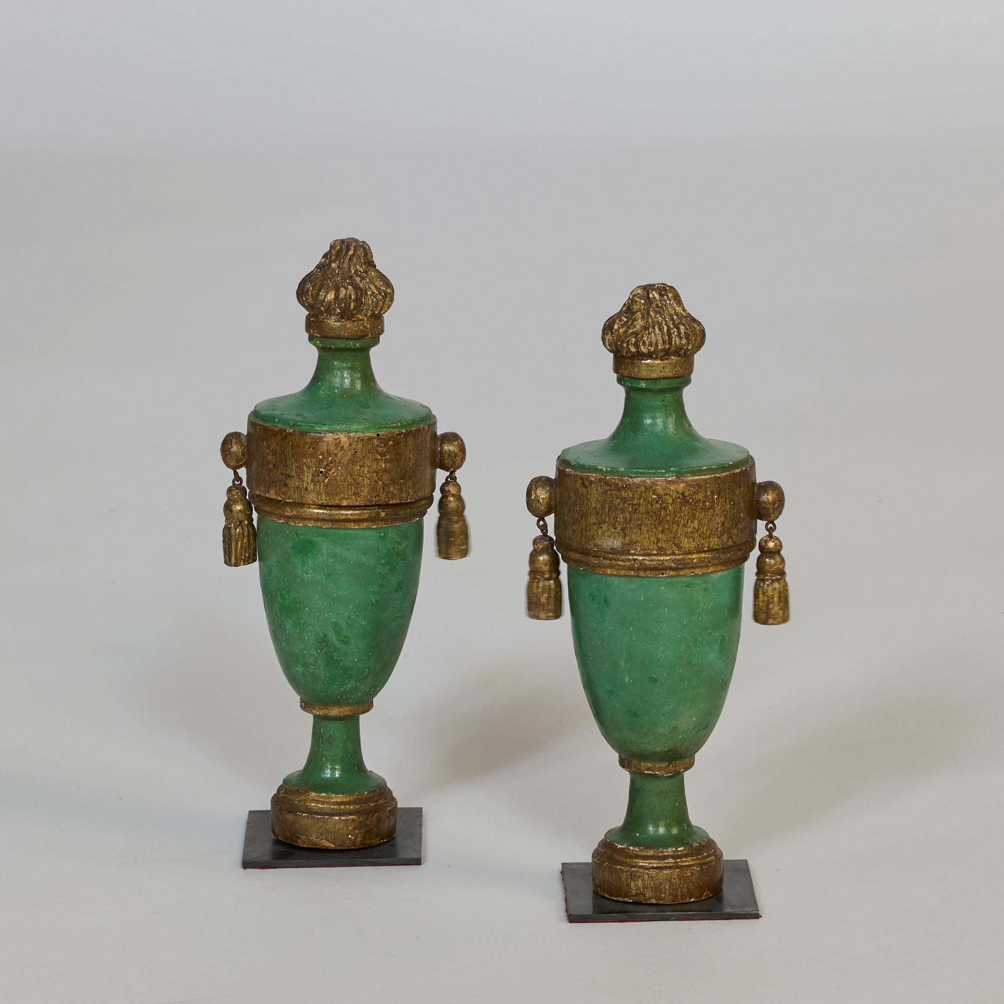 The Pair of Antique Style Green & Gilt Urns feature green bodies with gold accents, lids, bases, and side tassels, all set atop square black bases—ideal for bringing classical European decor to your space.