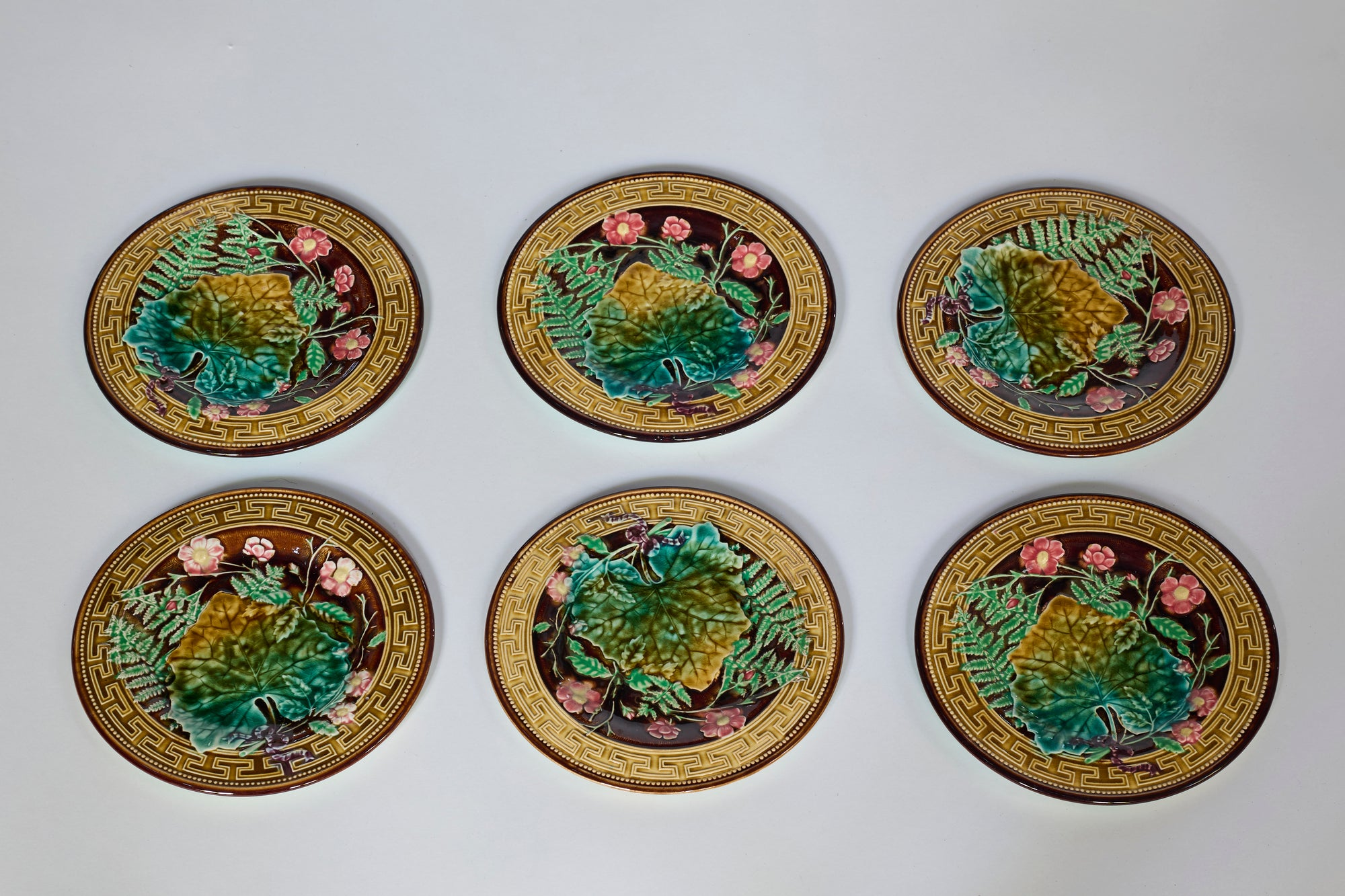 A Set of Six Antique Majolica Botanical Plates with Greek Key Border, showcasing green leaves, pink flowers, and yellow accents, are arranged in two rows of three on a plain white background.