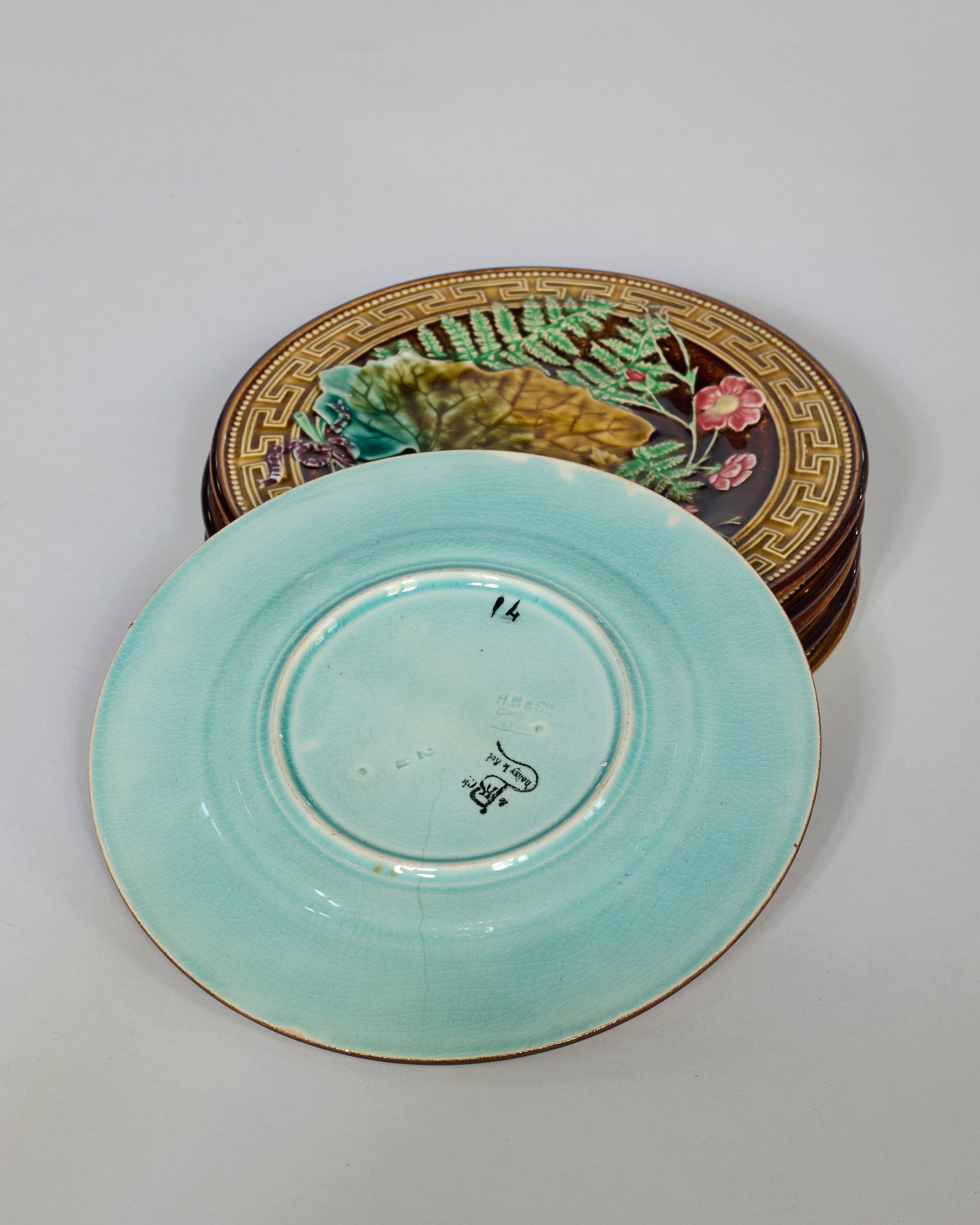 Set of Six Antique Majolica Botanical Plates with Greek Key Border, featuring colorful botanical motifs and geometric patterns. One plate is shown flipped, displaying its turquoise underside and visible maker’s marks.