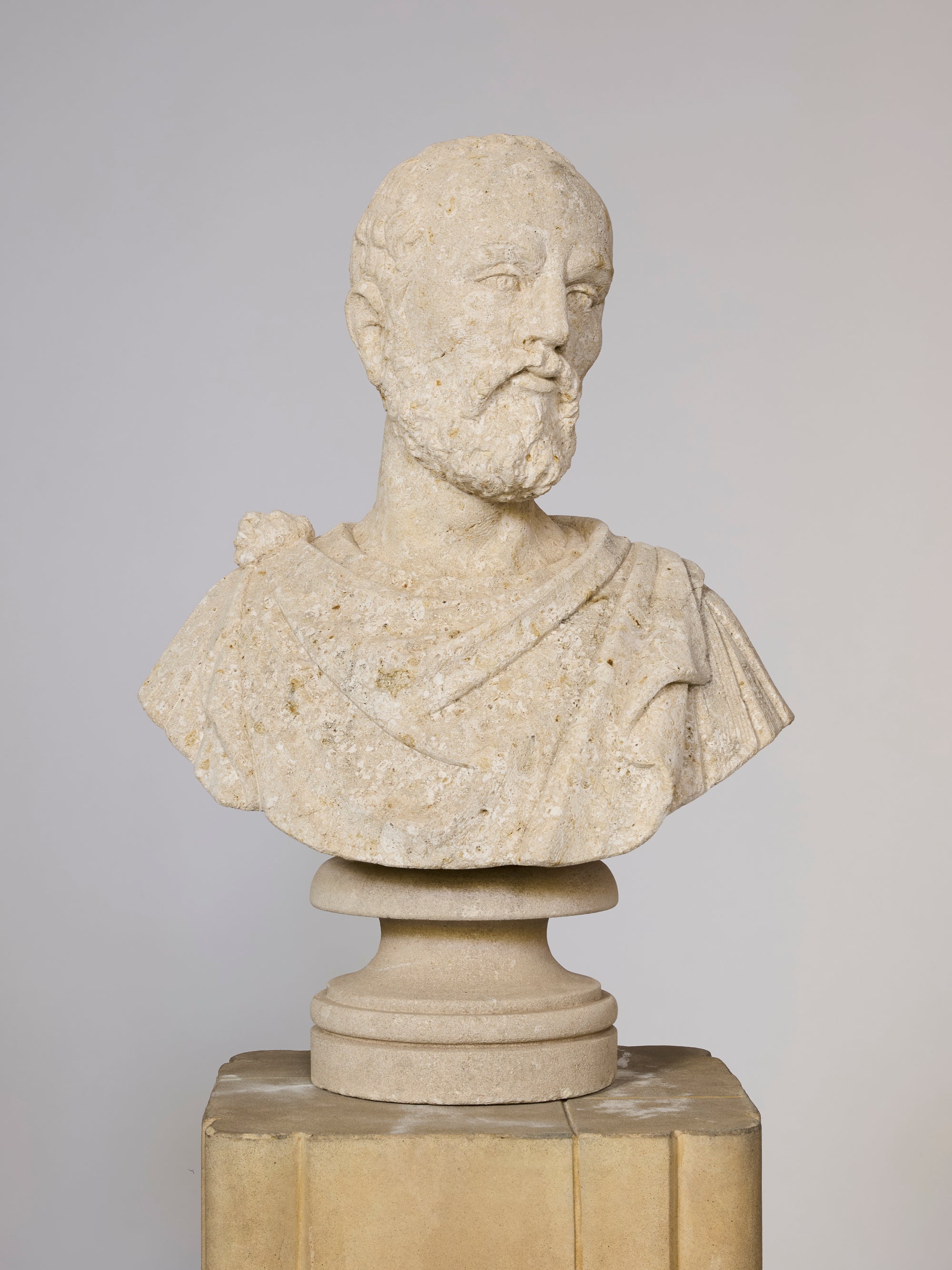 The Classical Style Coral Bust of a Bearded Philosopher features a short-haired, bearded man in draped attire on a pedestal, capturing the refined elegance of Greco-Roman sculpture against a simple, light backdrop.