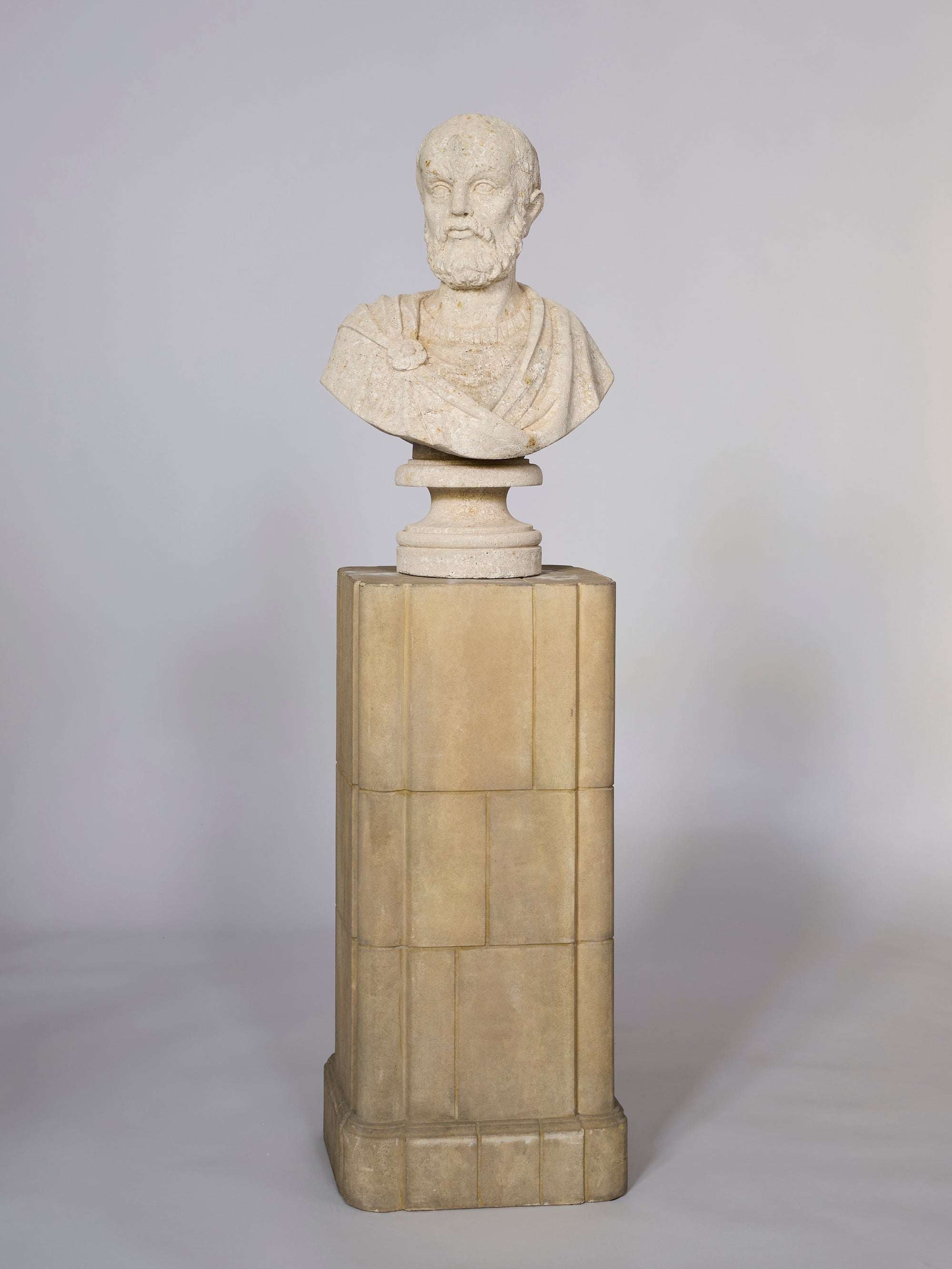 The Classical Coral Philosopher Bust features an elderly bearded man in a draped garment, set on a tall rectangular stone pedestal and displayed against a plain, light background.