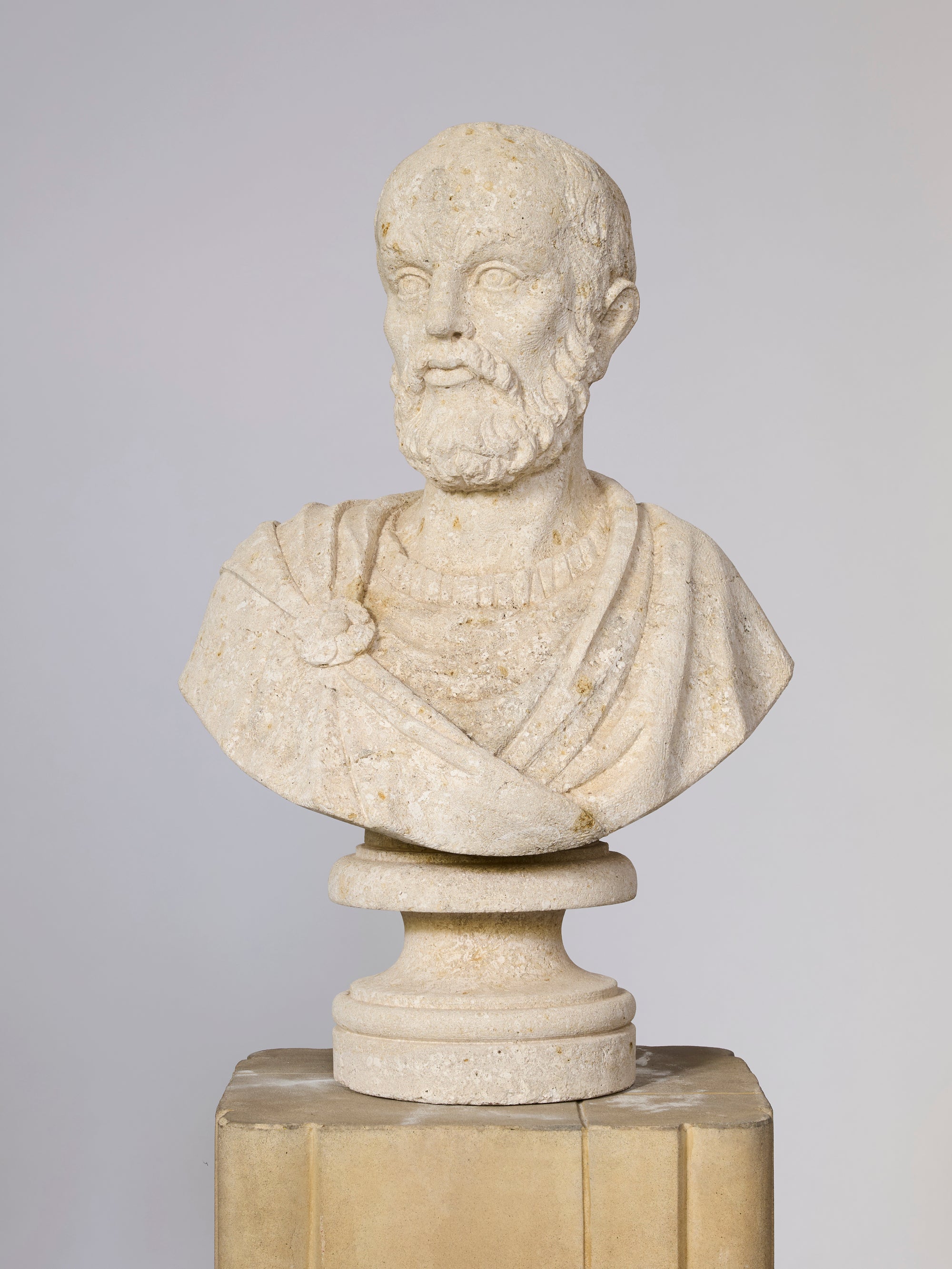 The Classical Coral Philosopher Bust, depicting a bearded man in Roman attire with a toga and shoulder brooch, stands on a pedestal against a plain background.