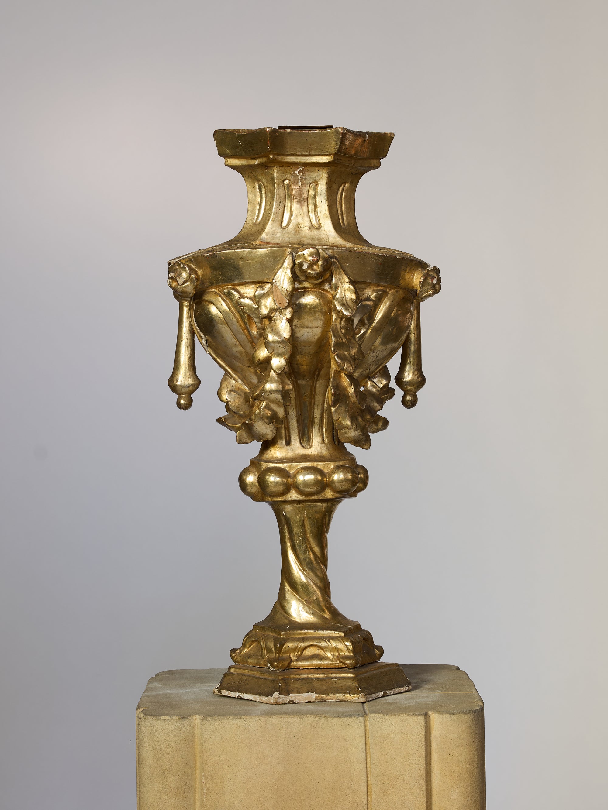 A Giltwood Classical Architectural Embellishment featuring figural and floral motifs, displayed on a beige pedestal against a plain light background.