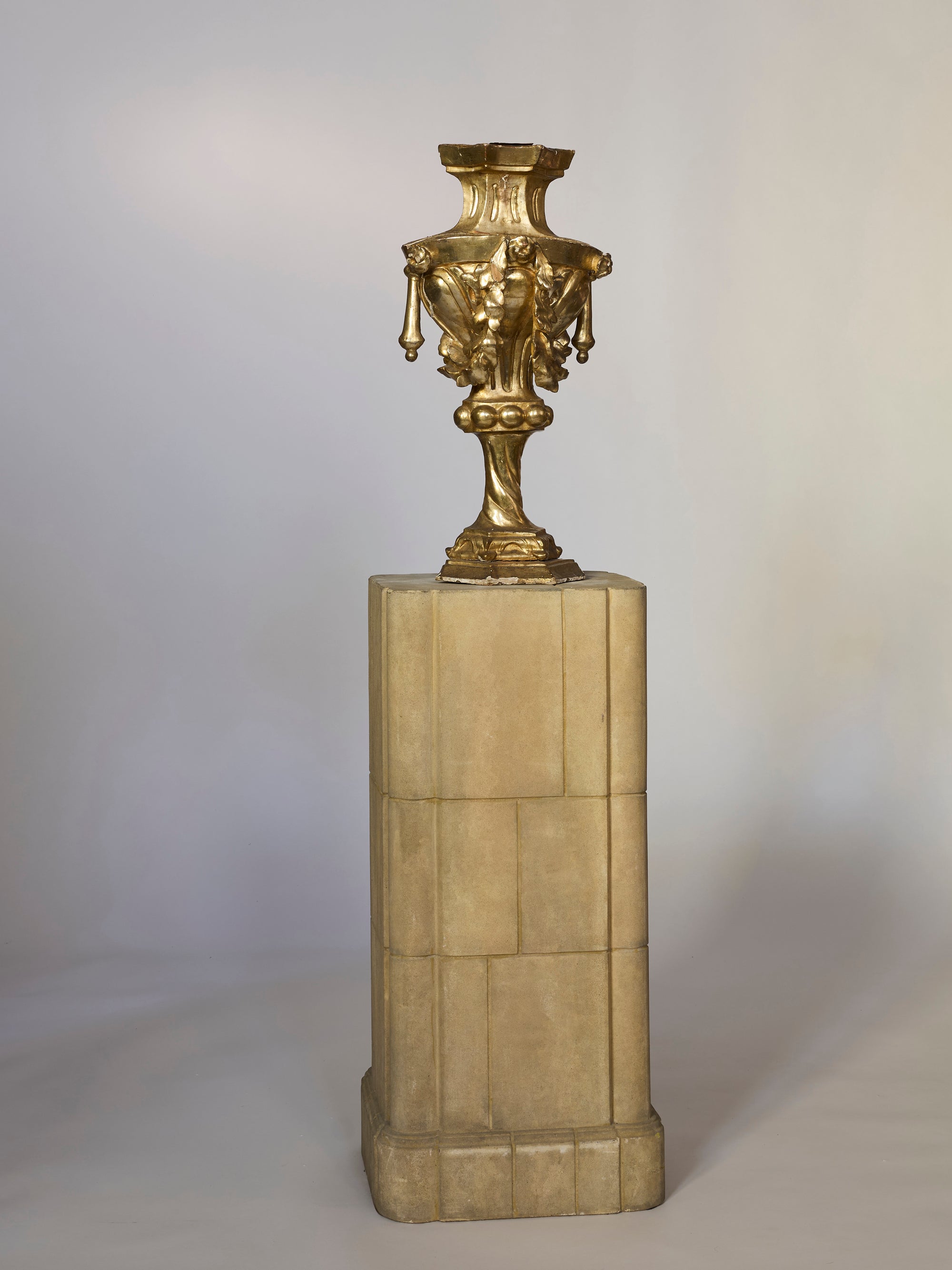 A Giltwood Classical Architectural Embellishment with figural and floral motifs, including ornate designs and decorative details, is displayed on a tall beige stone pedestal set against a simple, light background.