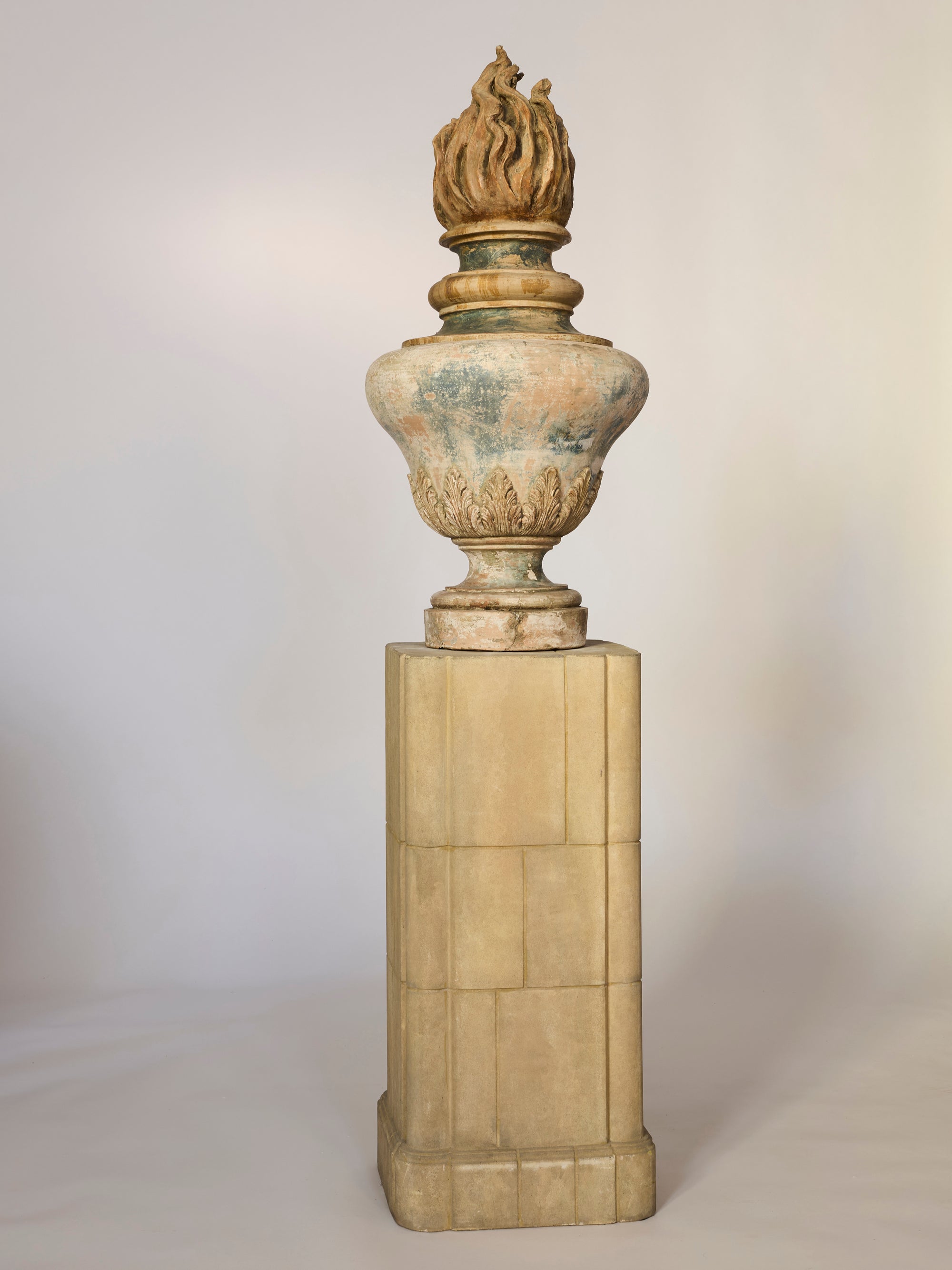 The Classical Flame Finial Urn Sculpture, featuring a weathered stone finish and classical flame finial, stands on a tall rectangular pedestal against a plain light background.