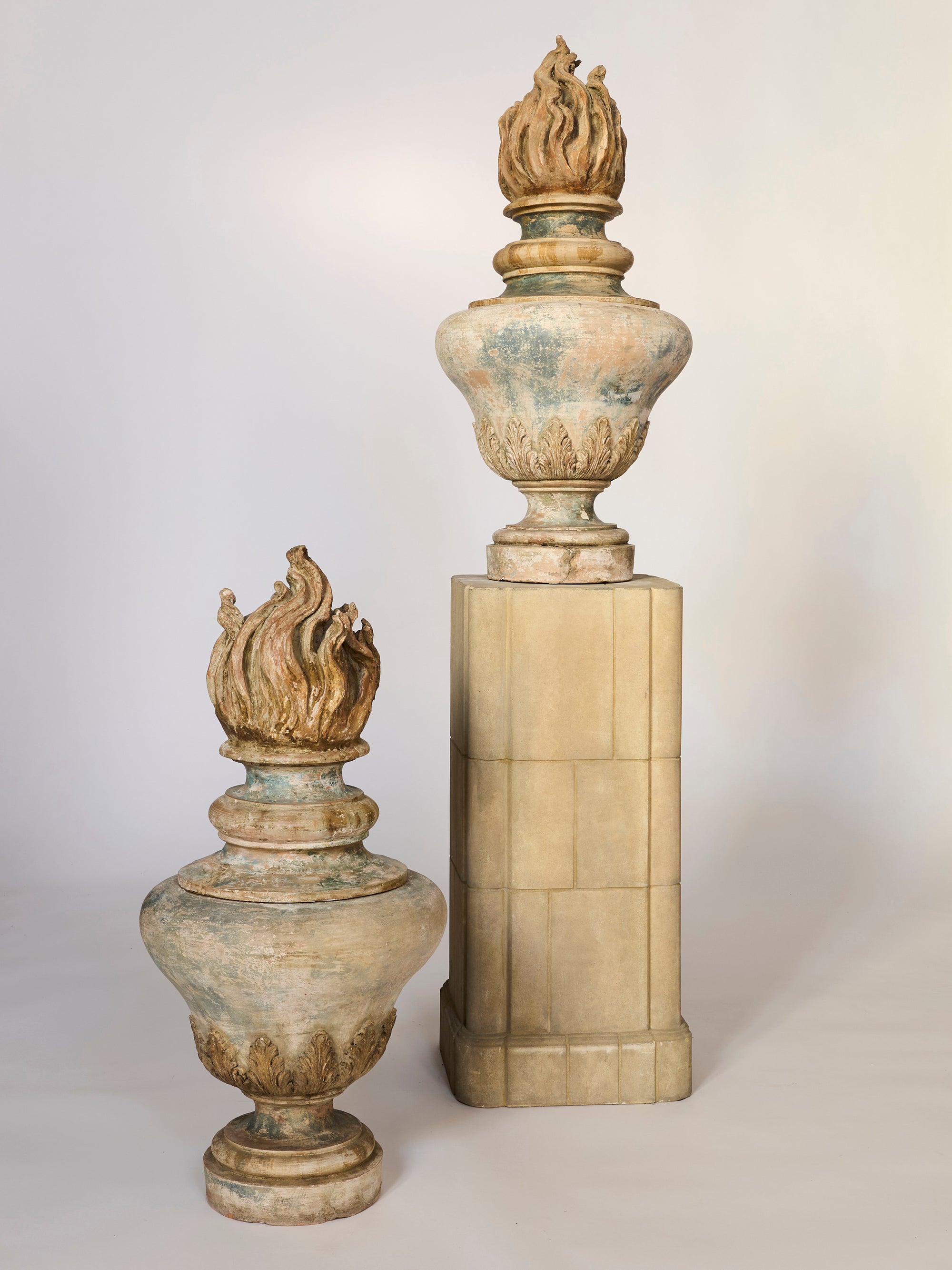 Two Classical Flame Finial Urn Sculptures—one grounded and one on a tall pedestal—feature ornate designs and weathered, aged finishes for an elegant architectural accent.