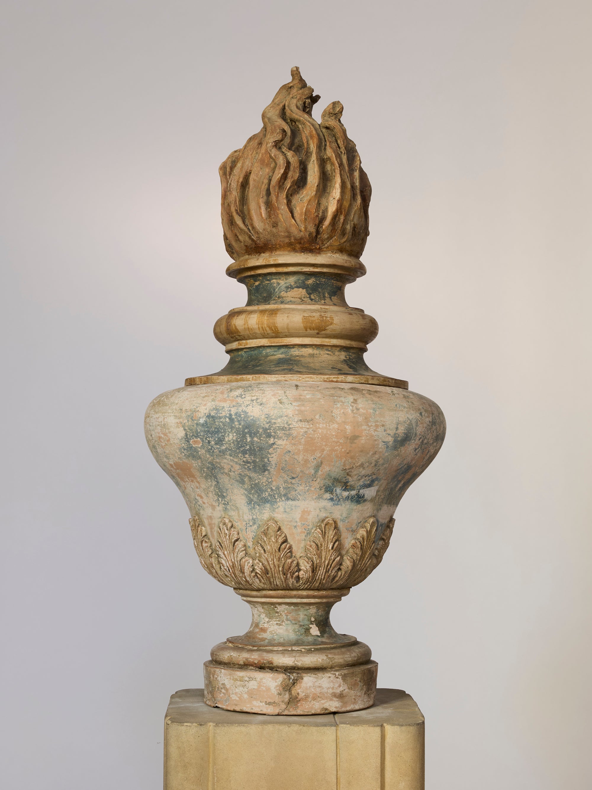 The Classical Flame Finial Urn Sculpture features carved leaf patterns, a flame finial, and weathered details that highlight its age, standing on a pedestal against a light background as an elegant decorative accent.