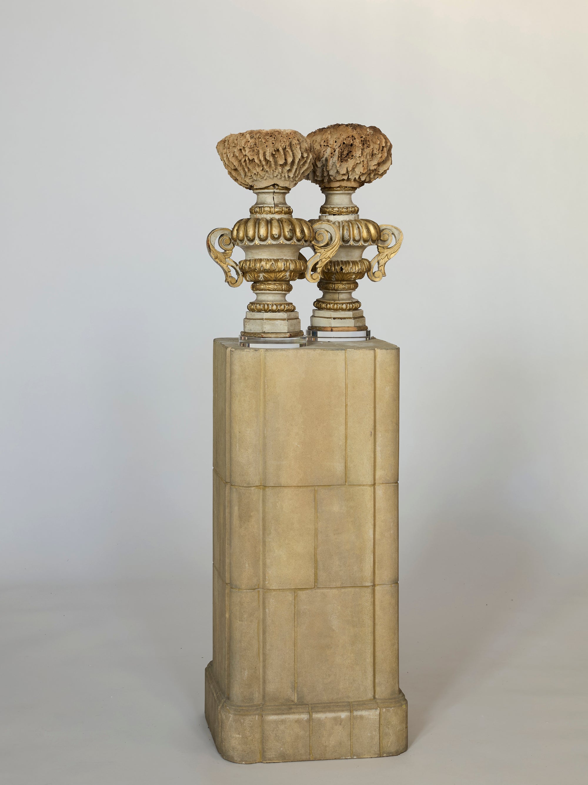 A Pair of Antique Architectural Urn Finials, featuring ornate gold and cream designs with textured tops, are displayed side by side on a tall stone pedestal against a plain light background.