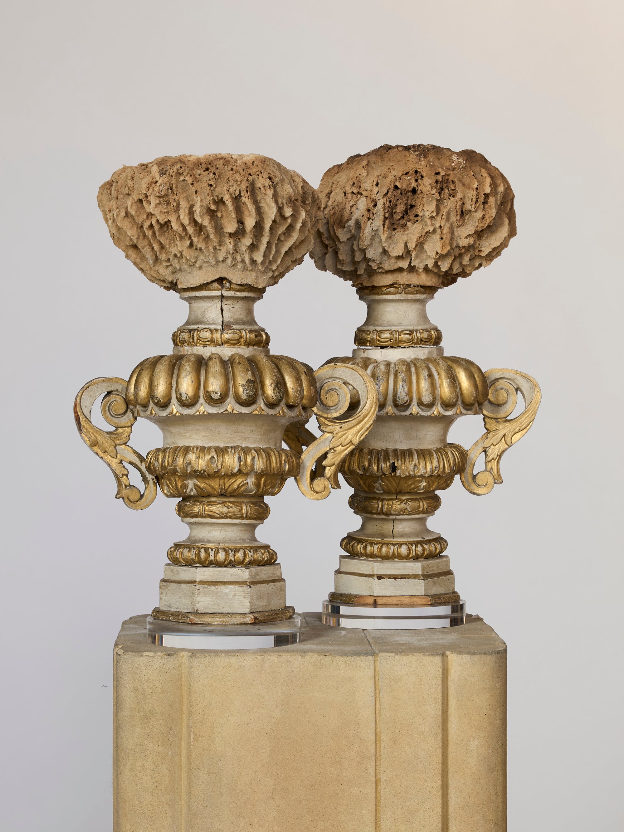 Two Antique Architectural Urn Finials in gold and cream with intricate handles are topped with brown, porous coral, displayed on a beige pedestal against a plain background.