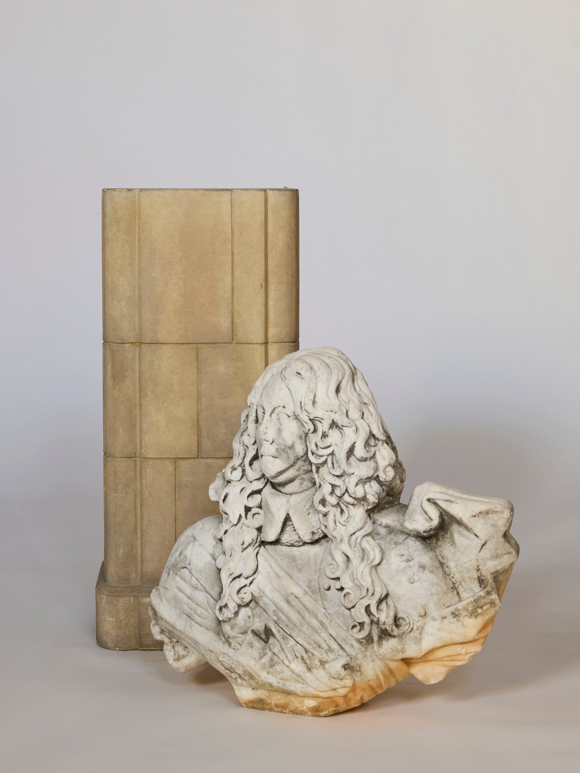 The Fragmentary Baroque-Style Stone Bust features a man with long, curly hair and ornate attire, positioned before a rectangular stone pillar on a neutral background. This aged sculpture displays visible signs of wear and partial damage.
