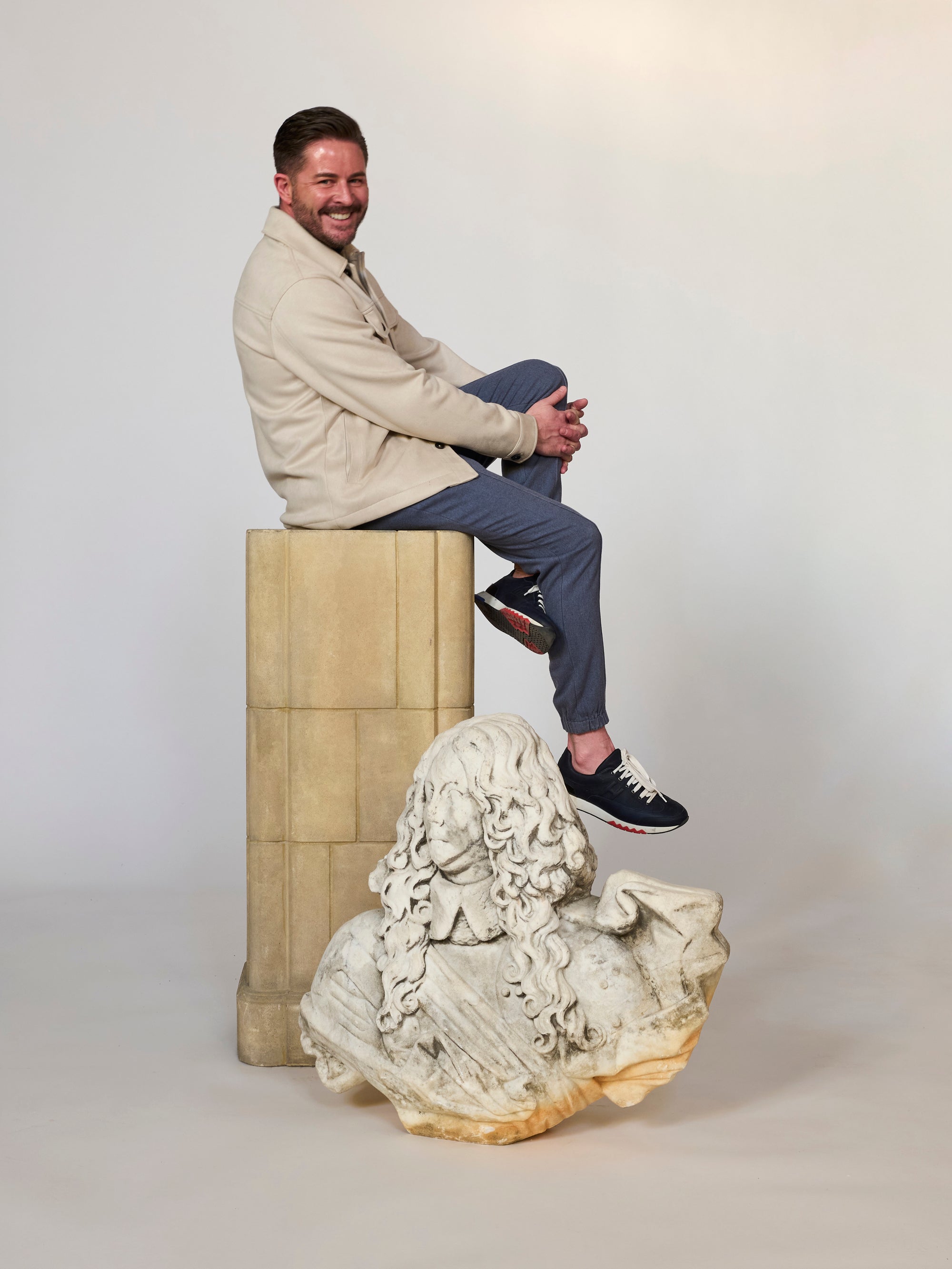 A man in casual attire sits smiling cross-legged on a square pedestal. In front of him, the Fragmentary Baroque-Style Stone Bust is displayed on the floor against a plain, neutral background.