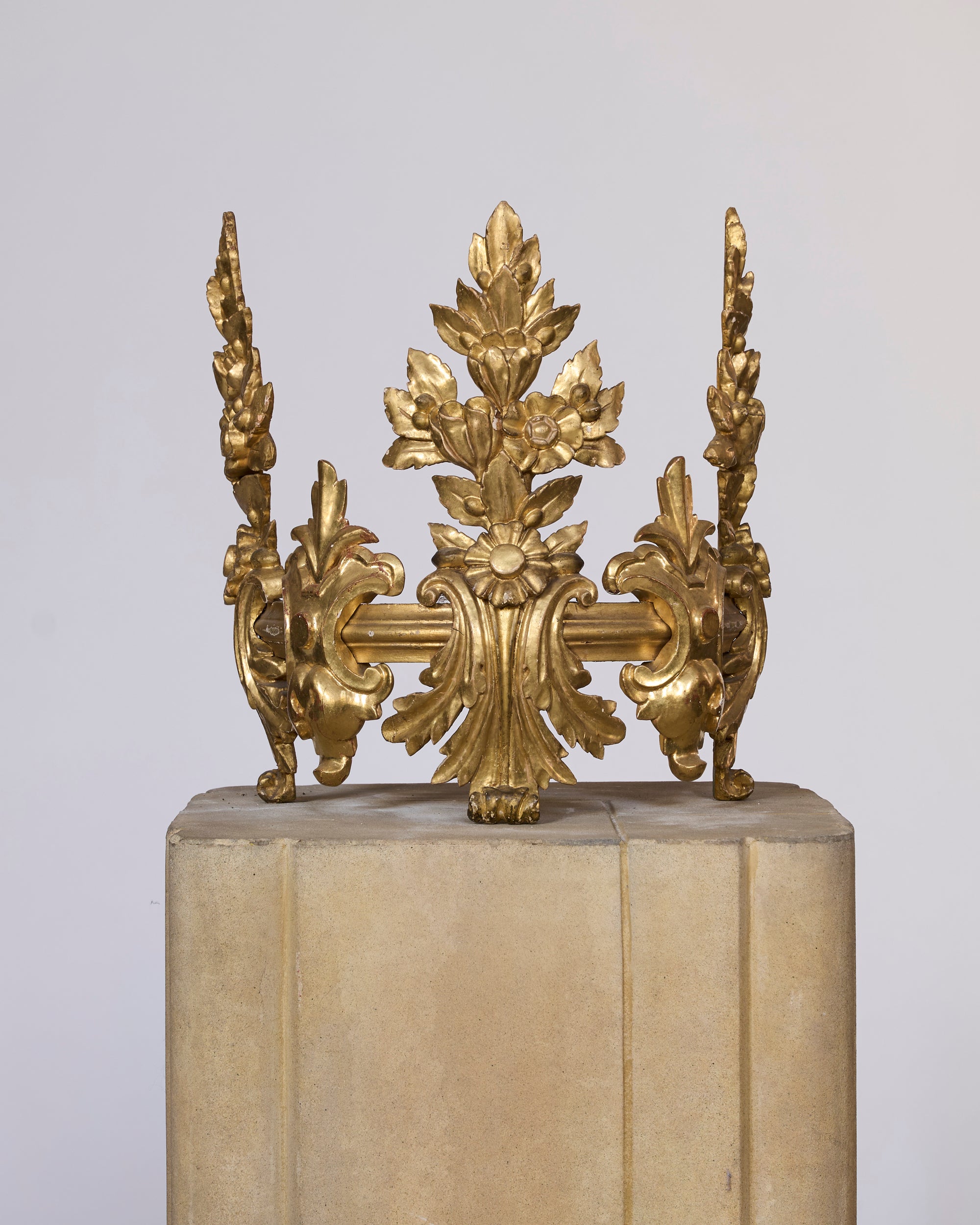 The Gilt Bronze Architectural Ornament, featuring ornate floral and leaf motifs inspired by classical European decor, is displayed atop a beige stone pedestal against a plain, light background.