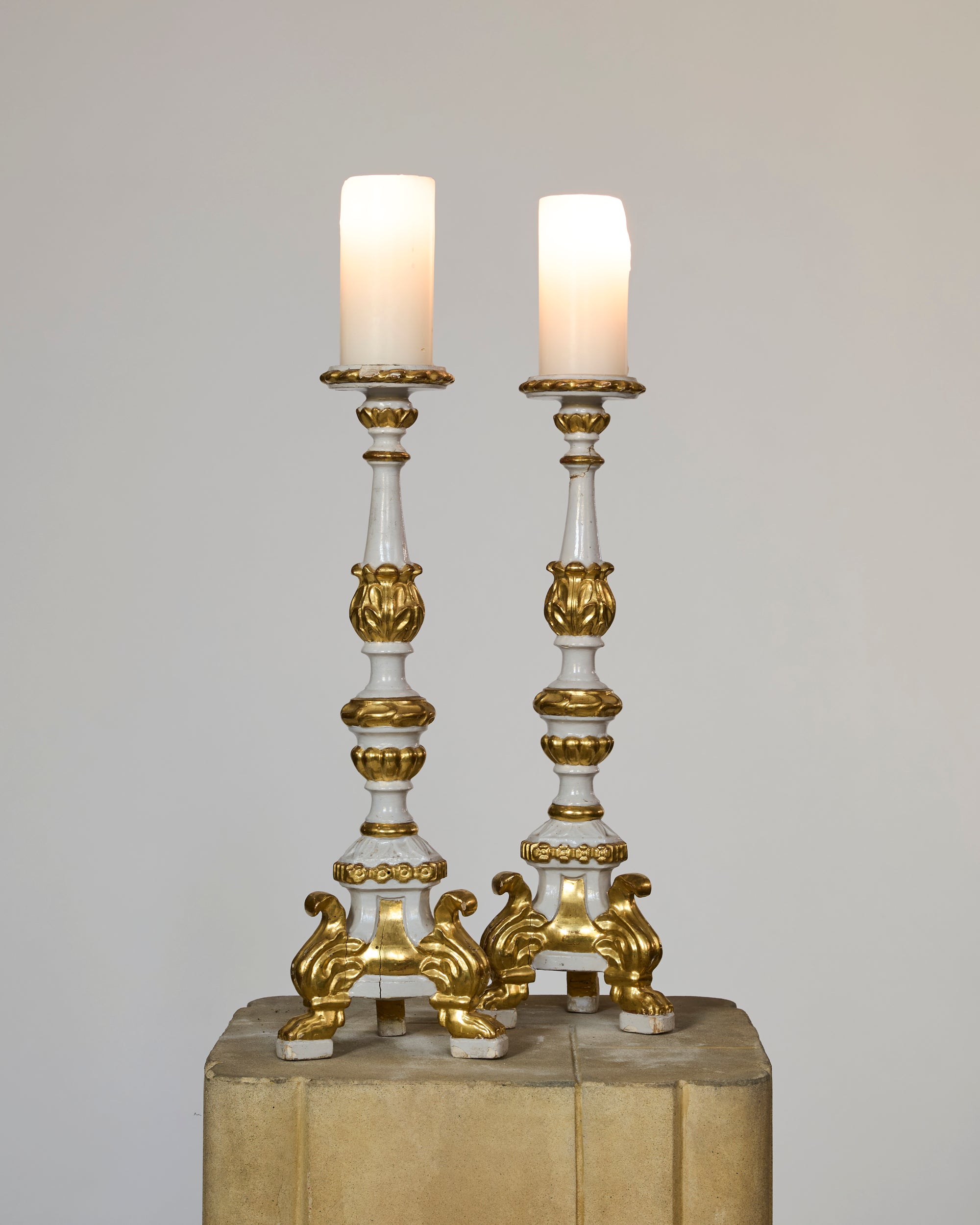 A Pair of Antique European Giltwood Candlesticks with White Painted Finish, each topped with a white pillar candle, stand side by side on a beige pedestal against a plain background.